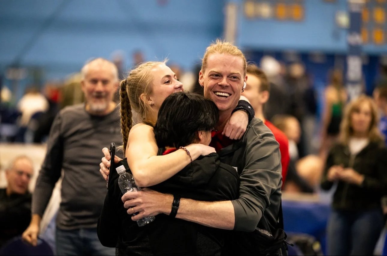 People hugging and smiling at an indoor event with a blue background and blurred crowd.