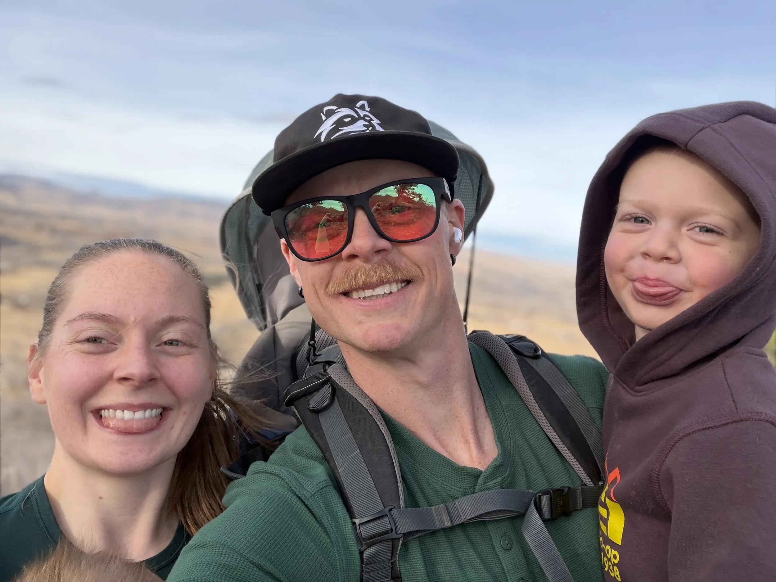 A family of four taking a selfie outdoors in a scenic landscape with hills and a blue sky. The father, wearing a black cap, sunglasses, and a backpack, is smiling. The mother, on the left, is also smiling with her tongue out. The young boy on the right has his tongue out and looks playful, wearing a brown hoodie.
