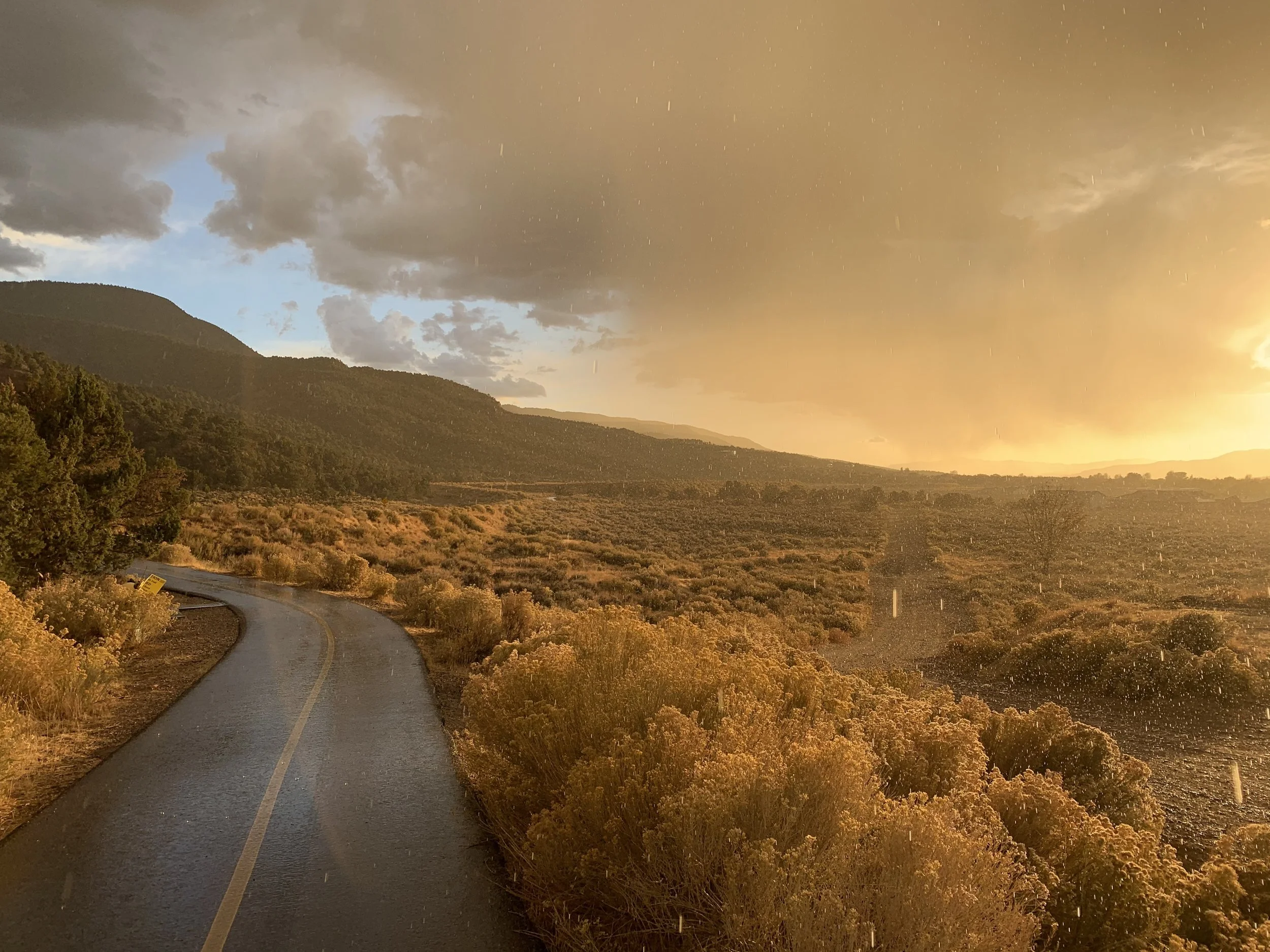 A winding rural road through a dry, hilly landscape with shrubs and bushes, under a dramatic sky with dark clouds and sunlight breaking through.