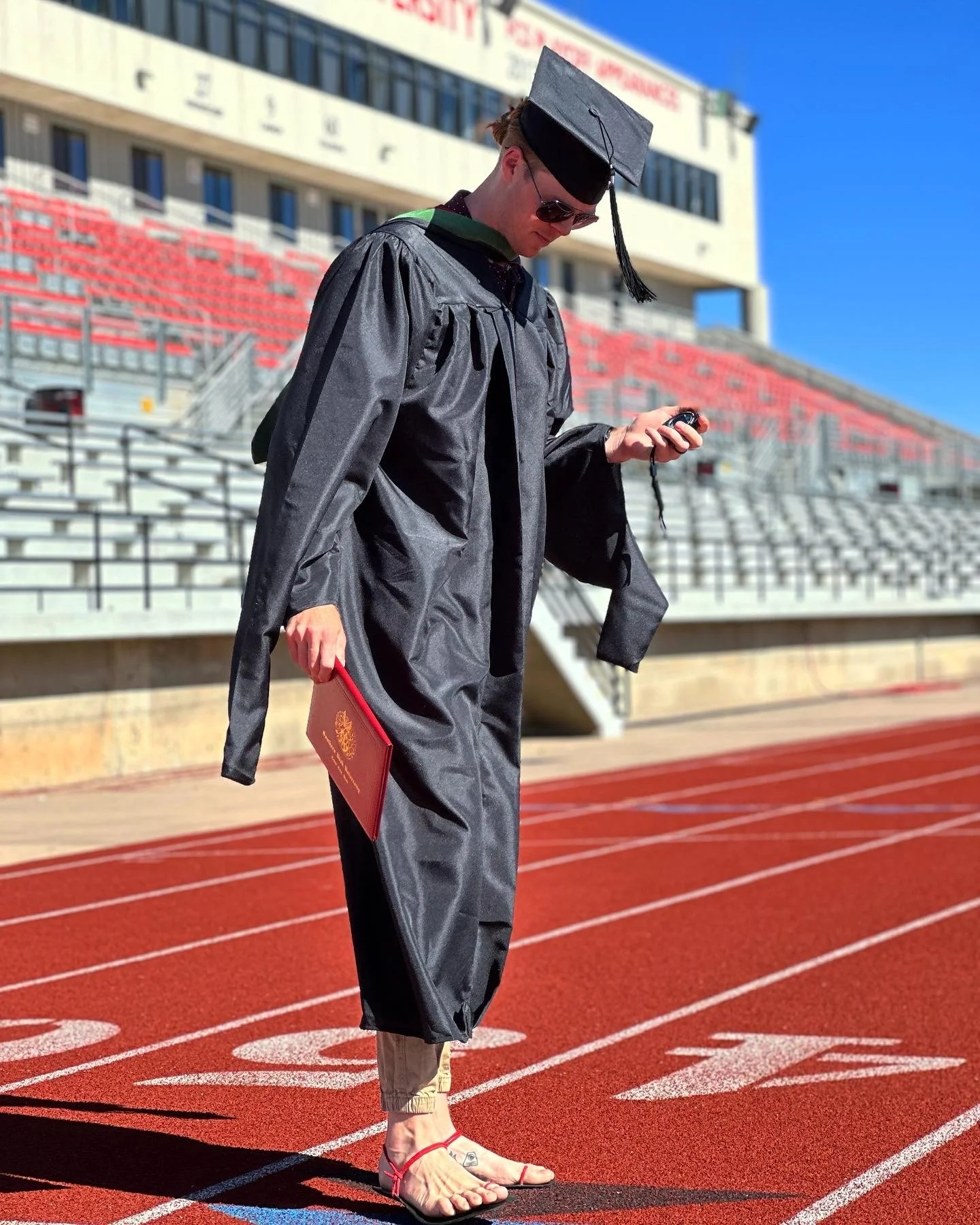 A graduate in academic regalia, holding a diploma, standing on a red running track at a stadium, looking at a phone, wearing sunglasses, and sandals.