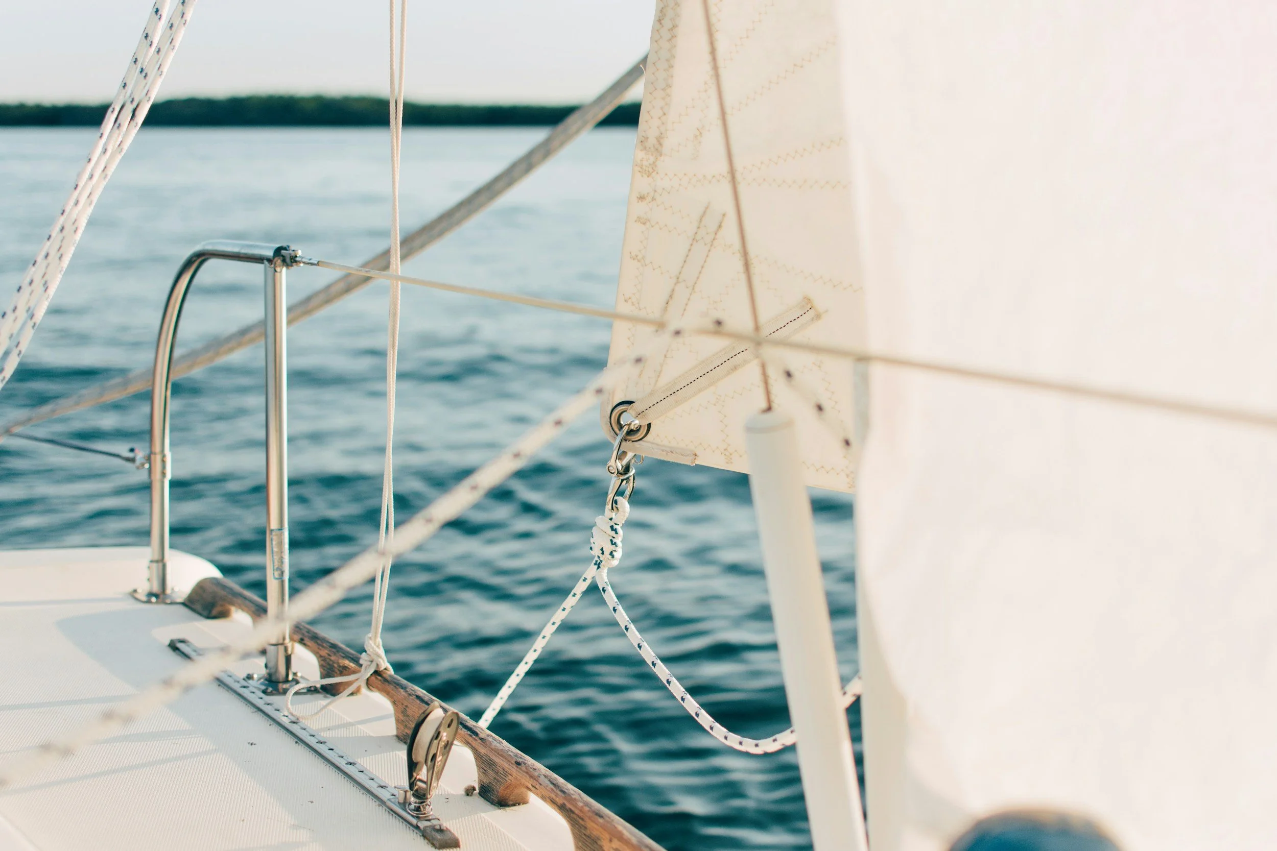 Close-up of a sailboat's rigging and a main sail on calm water with a distant shoreline in the background.