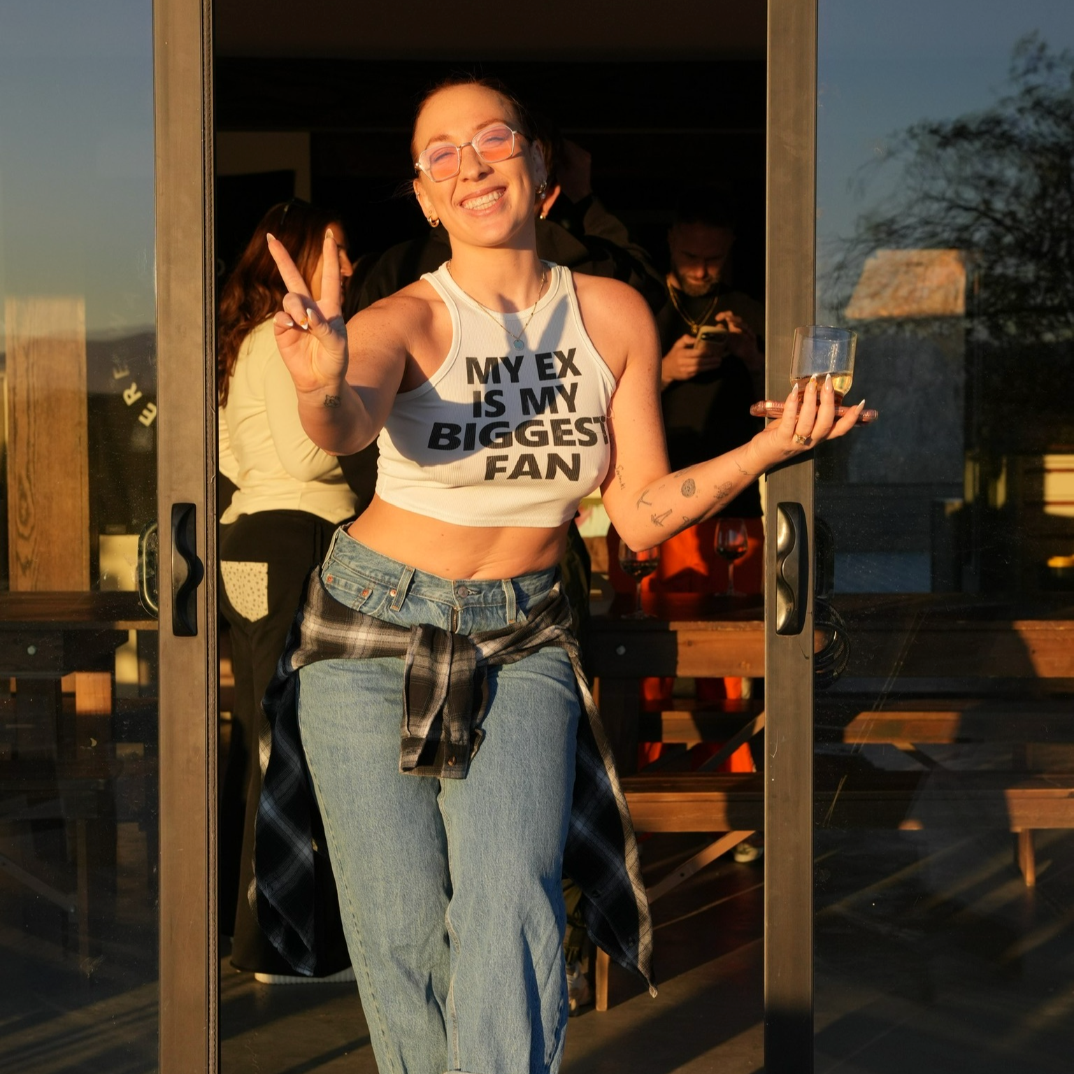 A smiling woman with glasses and tattoos, wearing a white tank top that says 'My ex is my biggest fan,' holding a drink, and making a peace sign at a doorway during sunset with people in the background.