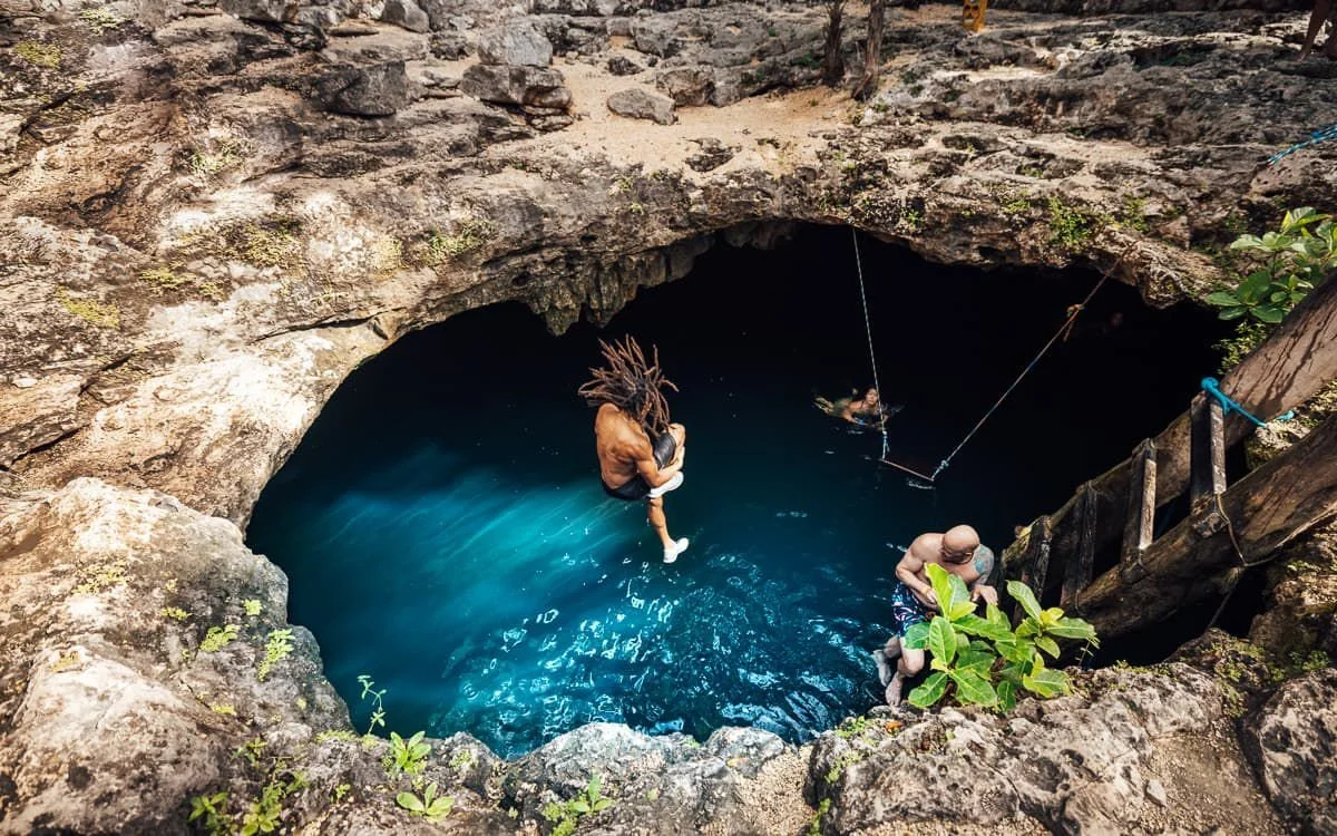 People swimming and exploring in a deep natural sinkhole or vertical cave with clear blue water, surrounded by rocky terrain and some greenery.