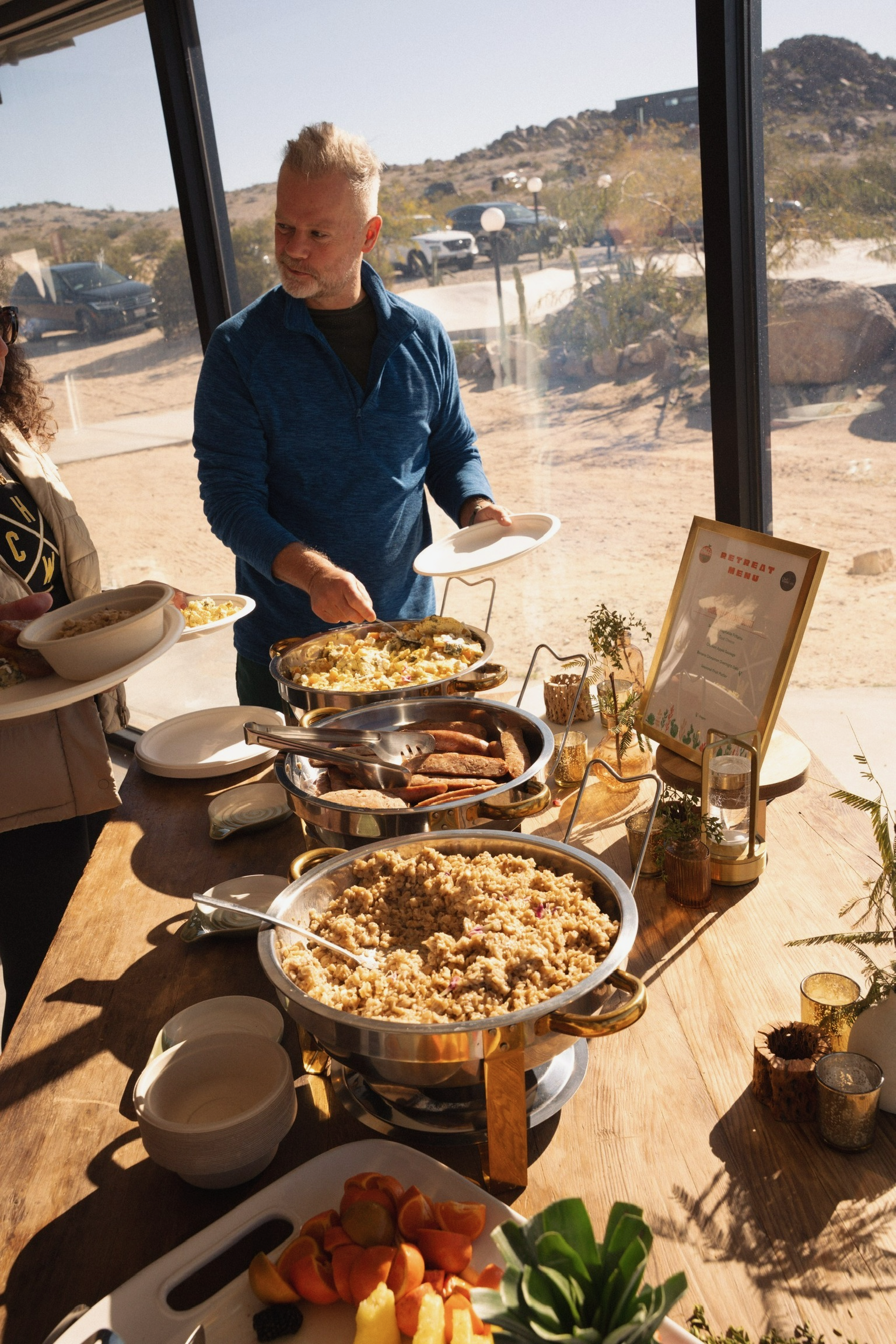 People serving themselves food at a buffet table with various dishes, including rice, sausage, and vegetables, inside a glass-walled room overlooking a desert landscape.