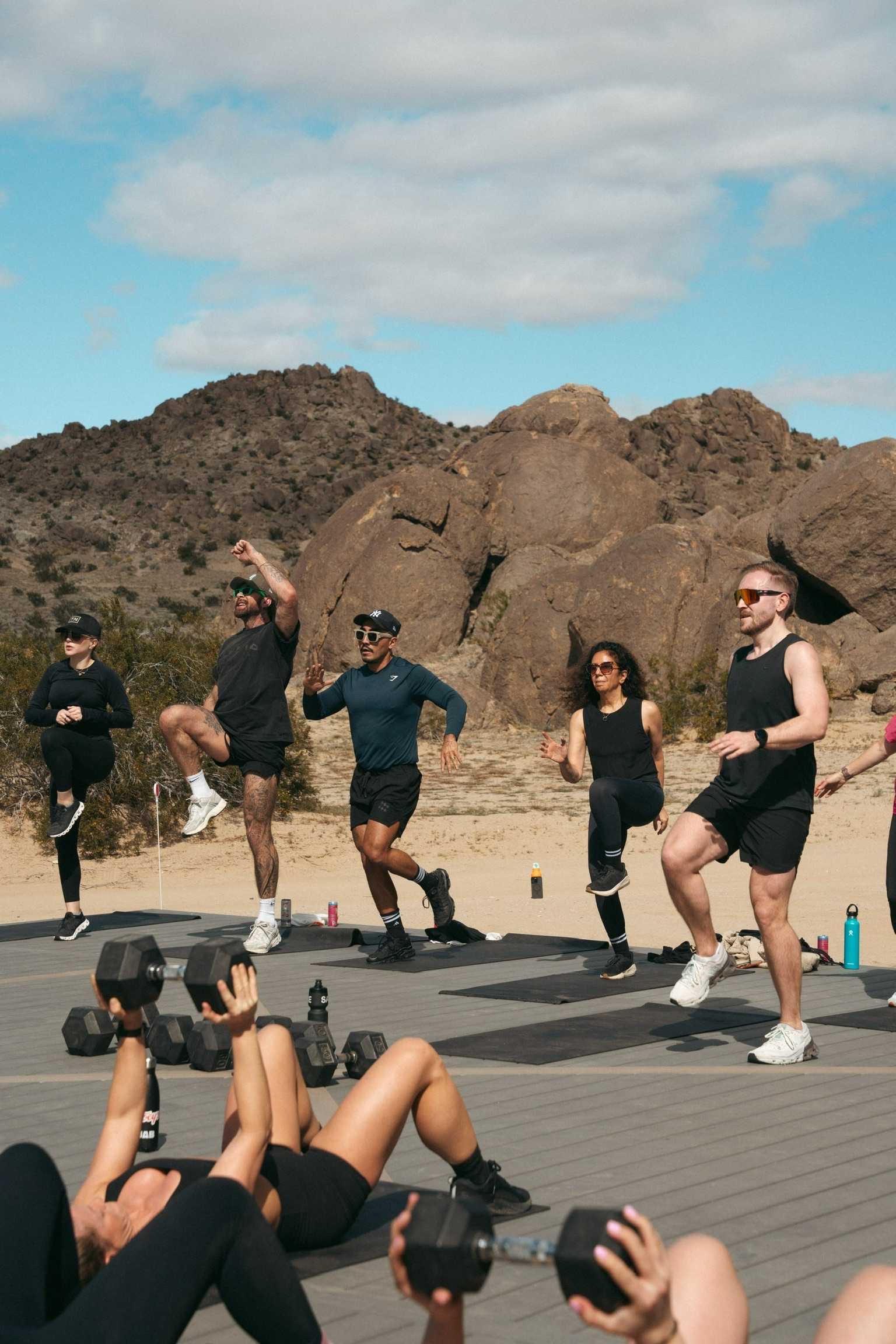 Group of people exercising outdoors on yoga mats in a desert landscape with rocks and mountains in the background. Some are standing on one leg, doing balance exercises, and others are lying on the ground lifting dumbbells.