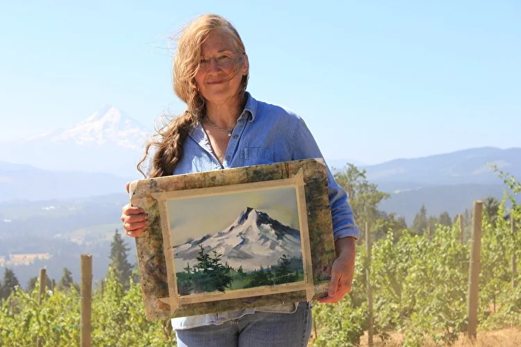 A woman holding a painting of a snow-covered mountain outdoors with a mountain in the background.
