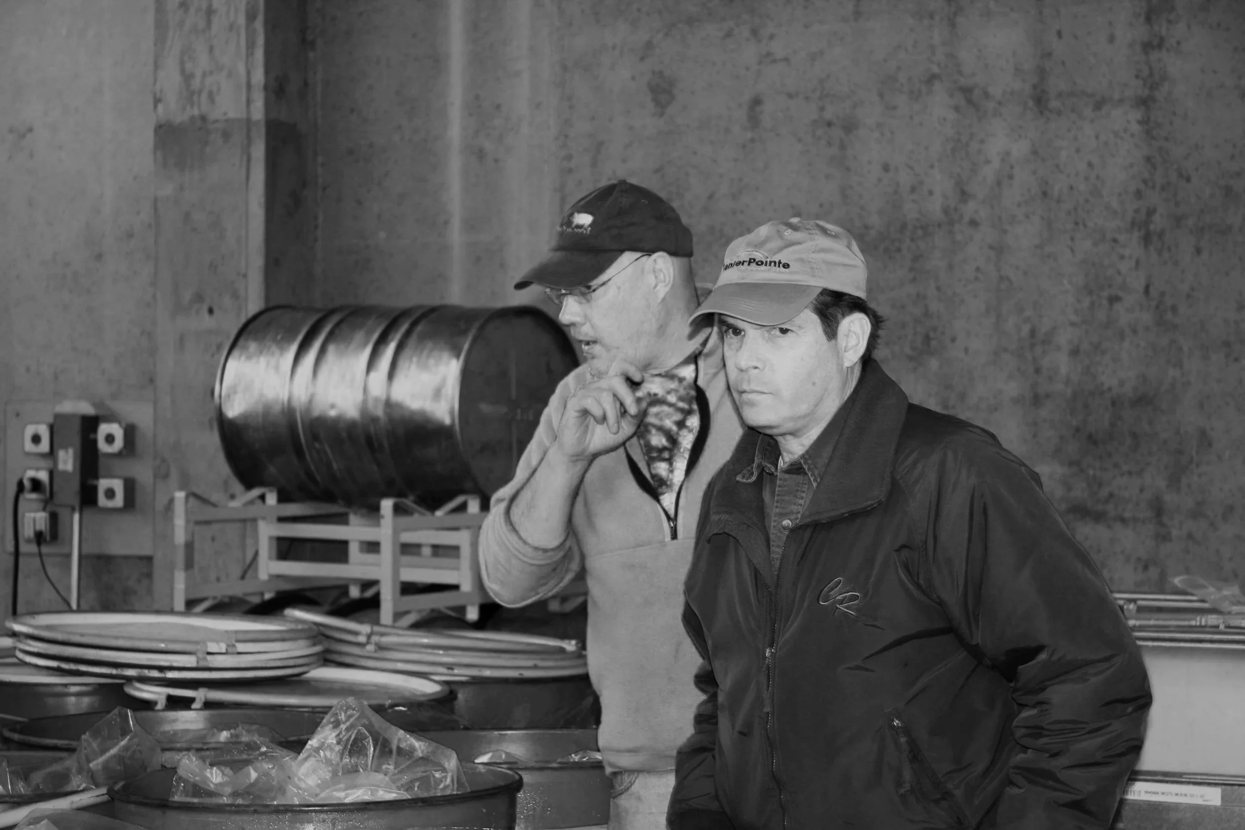 Two men in a warehouse or workshop, one in glasses and a cap, the other in a hat and jacket, standing near a table with various objects and a large cylindrical tank in the background.