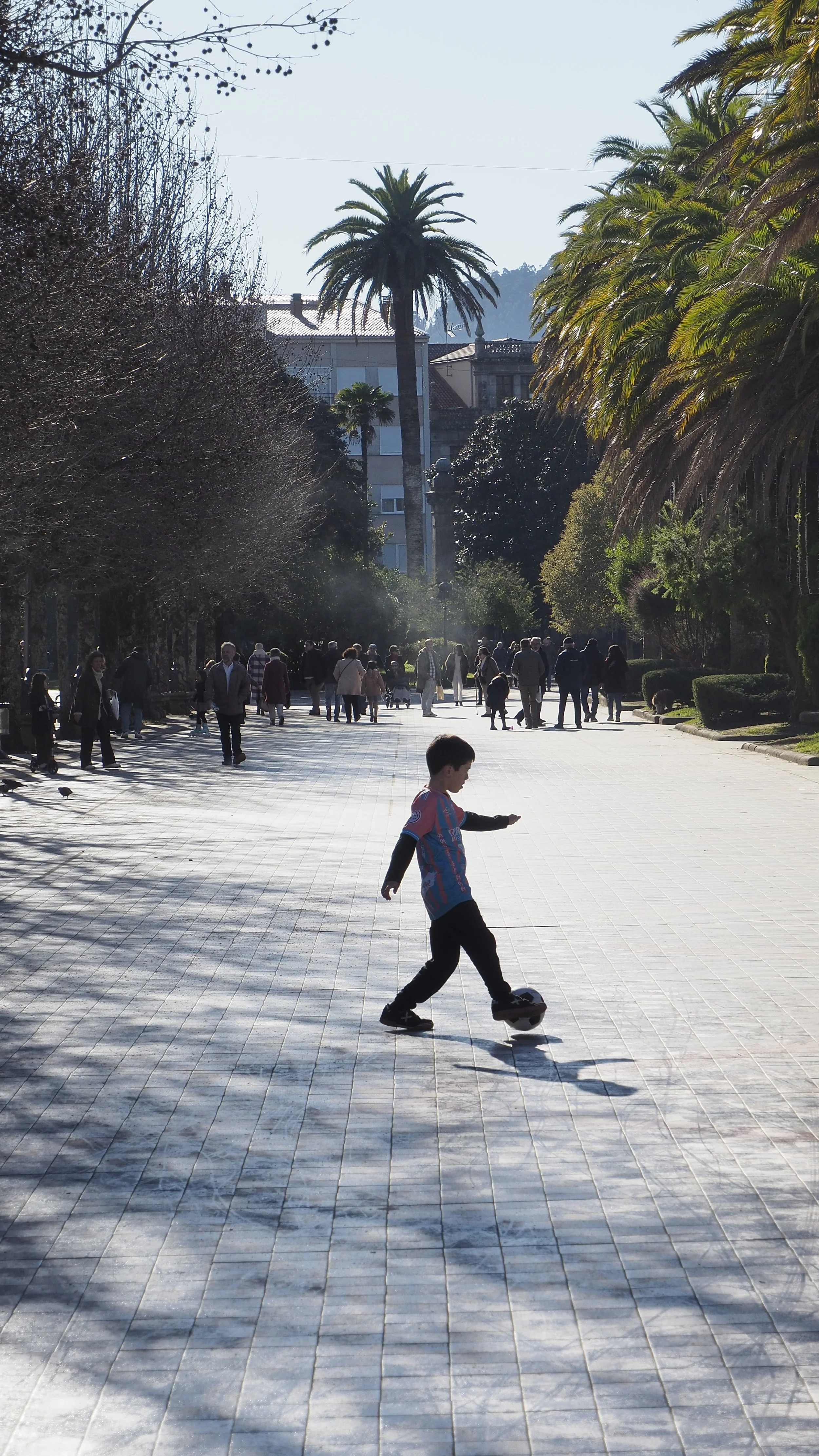 A young boy in a colorful sports jersey and dark pants playing soccer on a wide paved walkway in a park with many trees, palm trees, and people walking in the background.