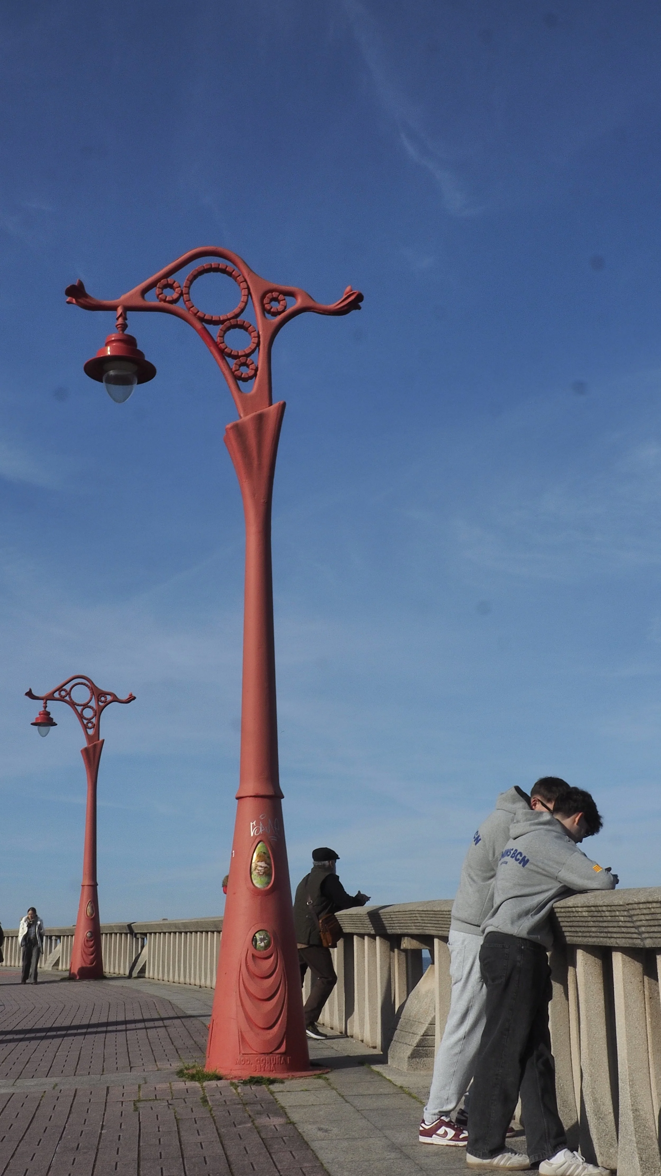 Two red decorative street lamps stand on a walkway, with three people leaning on a stone railing, looking over the water. The sky is blue with some wispy clouds.