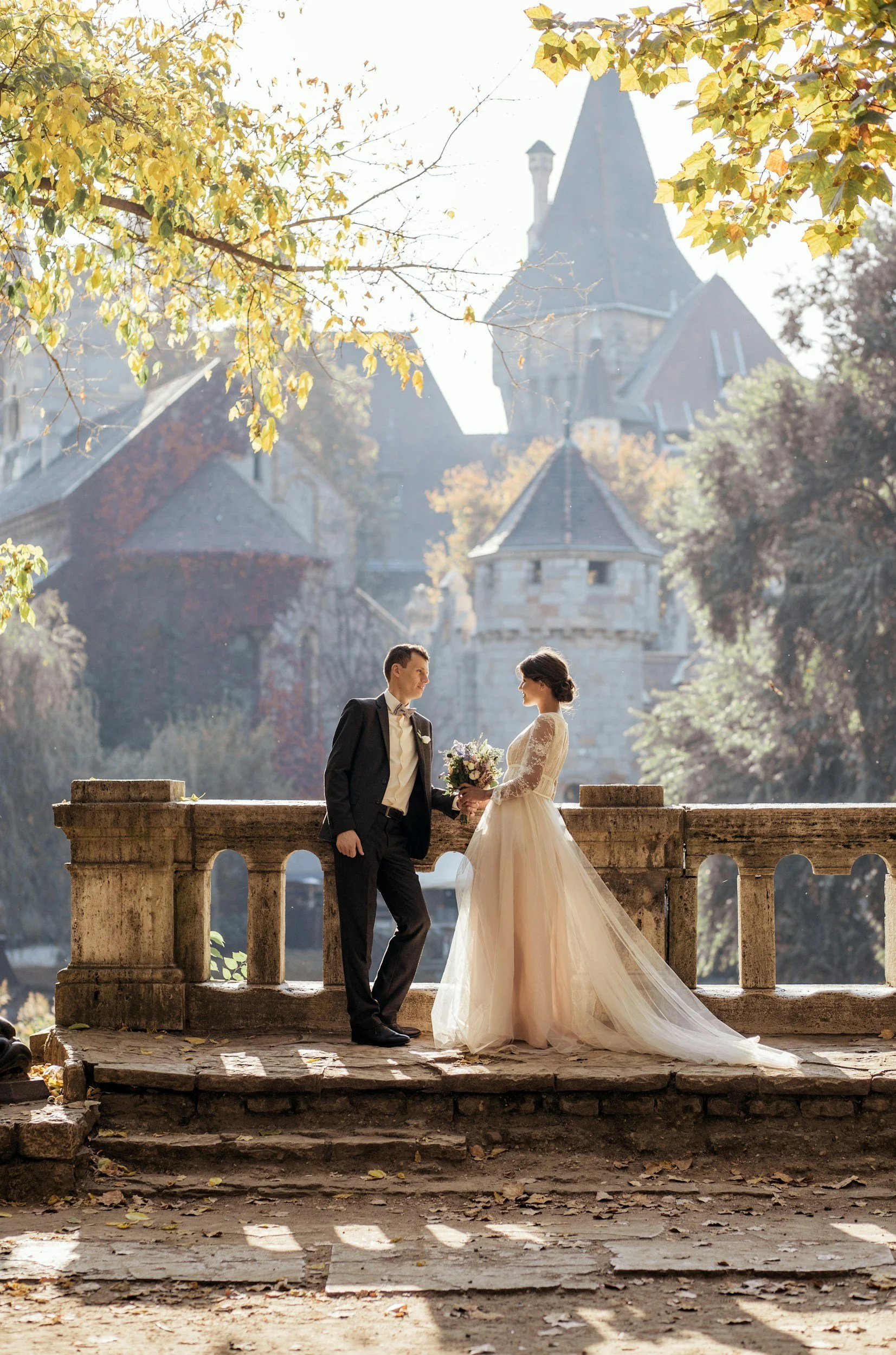 A bride and groom stand on a stone bridge, holding hands, with a castle-like building in the background. The bride wears an elegant, long-sleeved wedding gown and holds a bouquet of flowers. The groom is dressed in a black tuxedo with a bow tie. The 