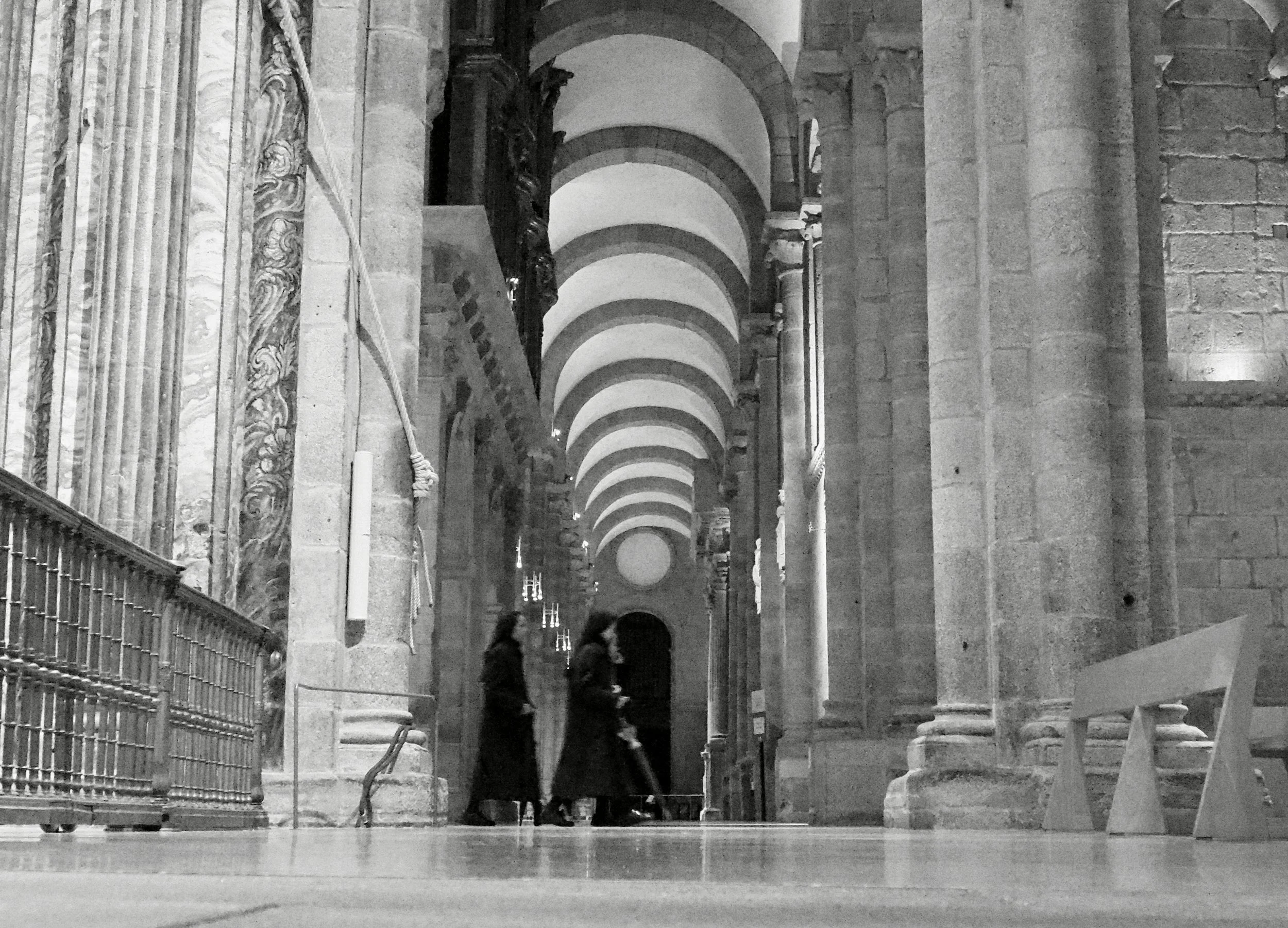 Interior view of a historic cathedral with high arched ceilings, stone pillars, and columns. Three women in dark clothing are walking and talking in the middle of the space. There is a small metal railing on the left and a bench on the right side.
