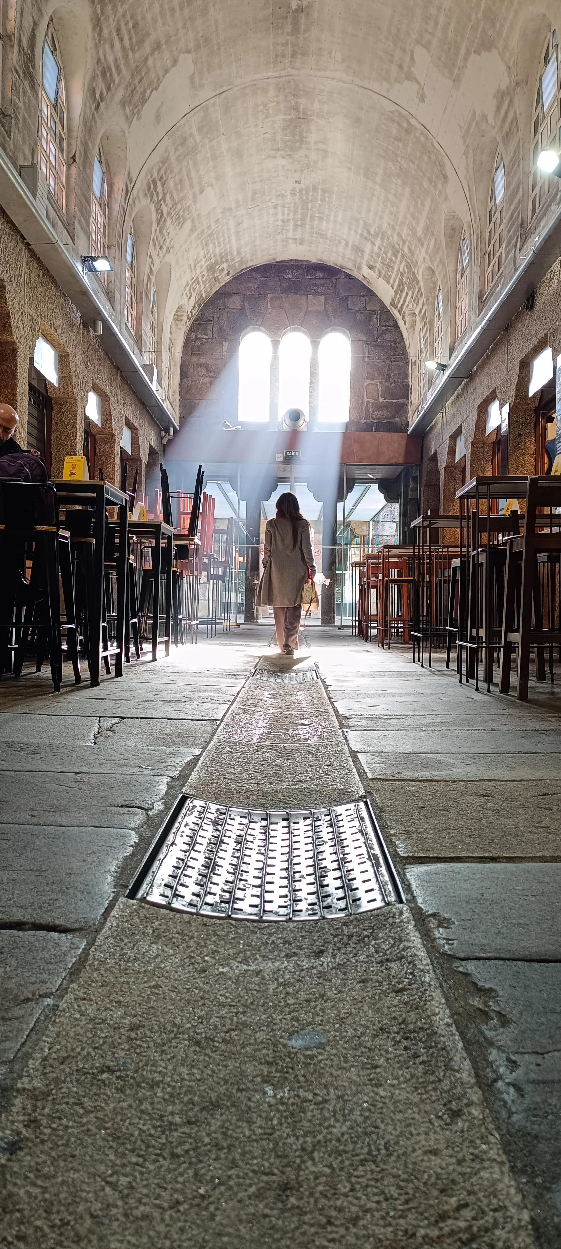 Interior of a historic building with a high arched ceiling, sunlight streaming through tall windows, and a woman walking toward the entrance.