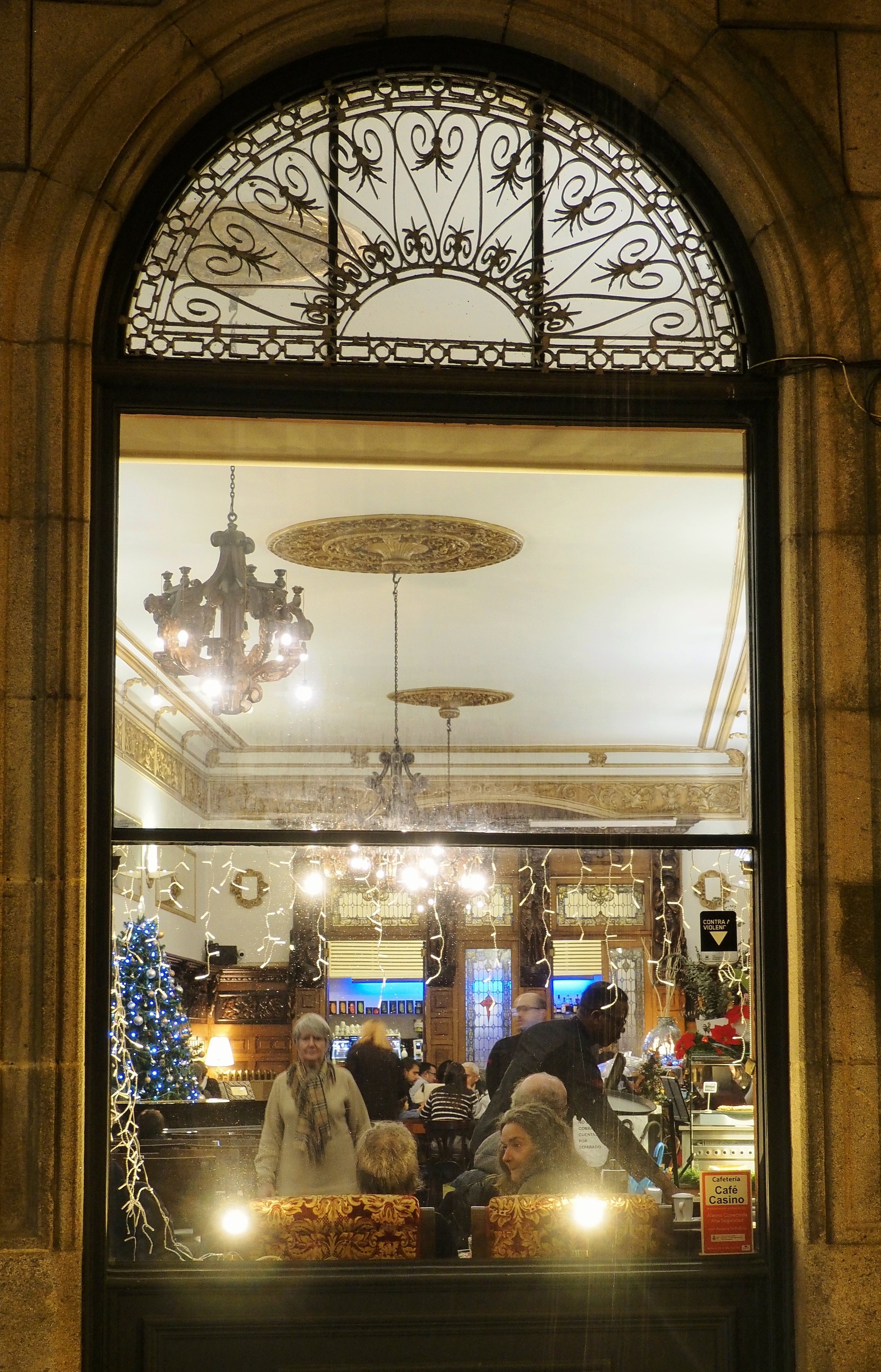 The image shows the interior of a decorated restaurant or cafe through a large window. Inside, there are elegant chandeliers, a decorated Christmas tree, and people dining. The room has ornate ceiling designs and wooden paneling, with string lights a