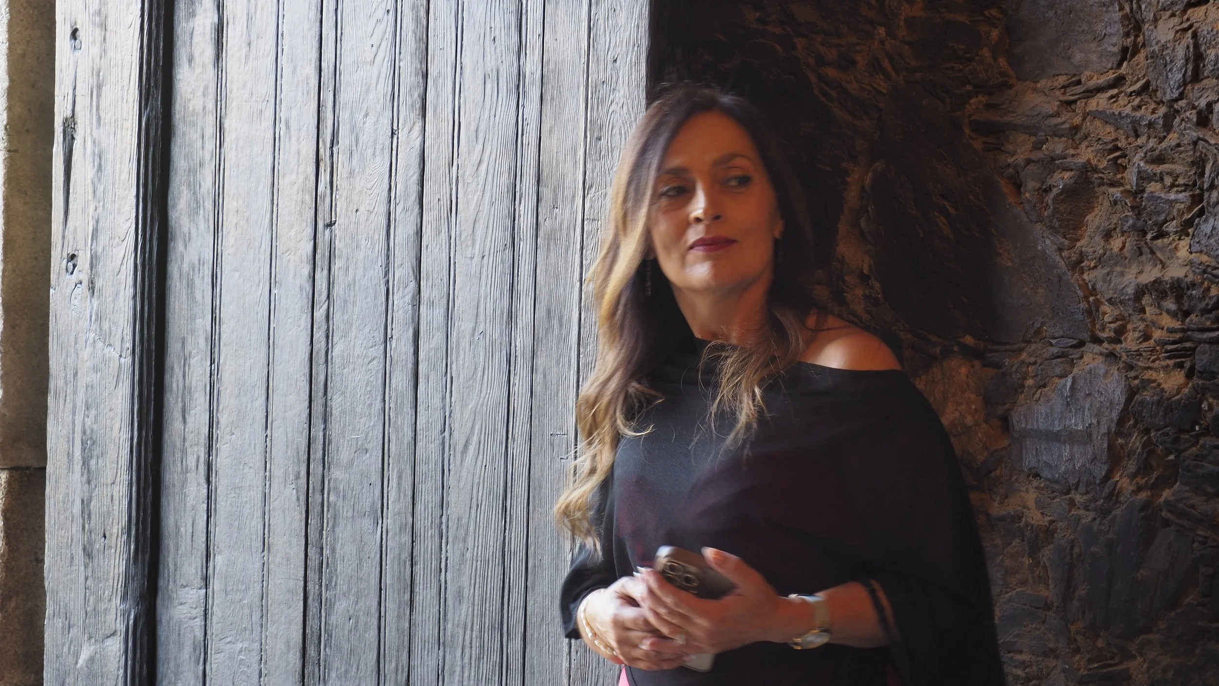 A woman with long, wavy hair wearing a black off-shoulder top, holding a phone in her right hand, standing against a textured stone wall next to a wooden door.