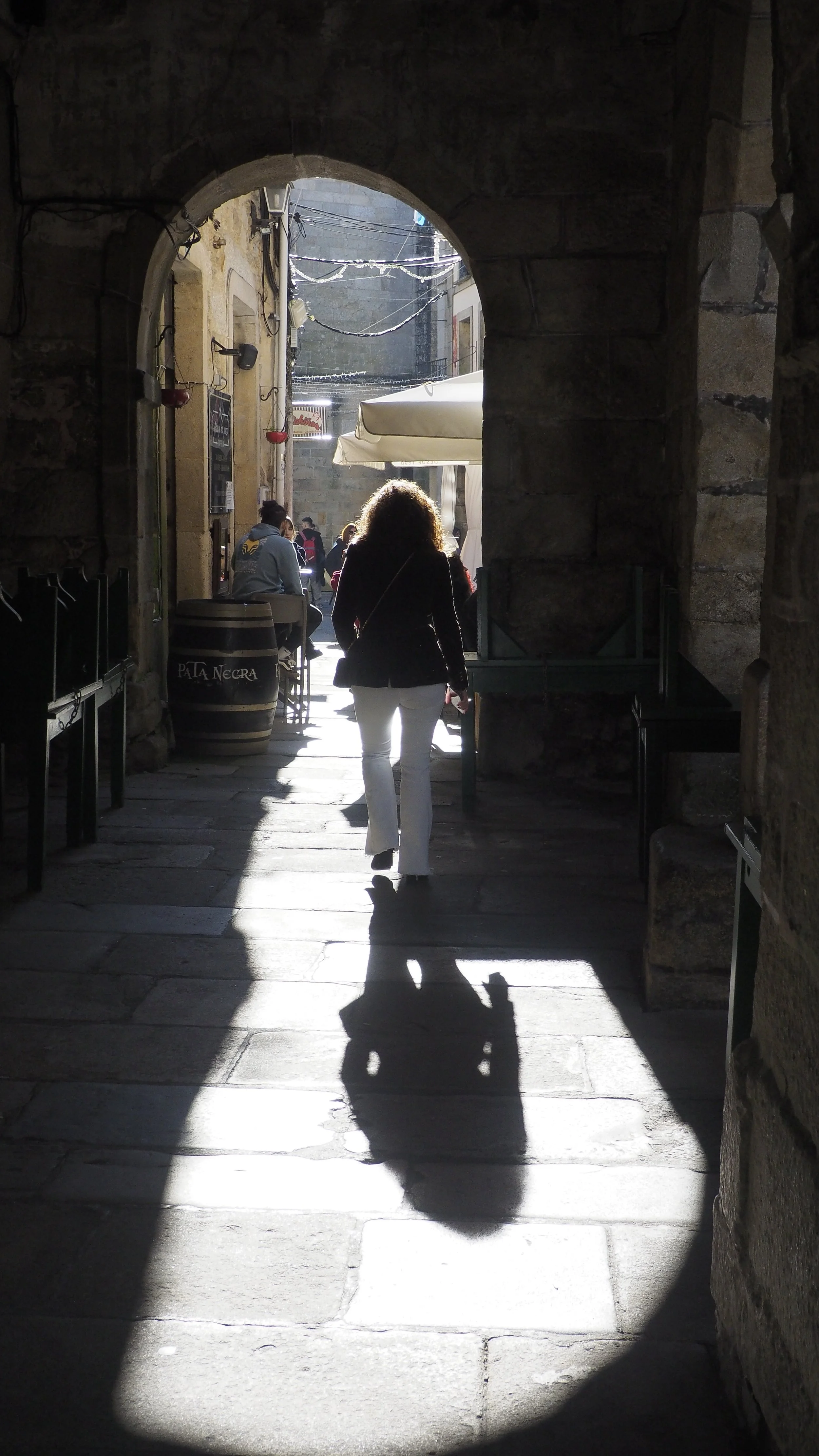 A woman walking through a stone archway into a sunny outdoor street with cafes and people.