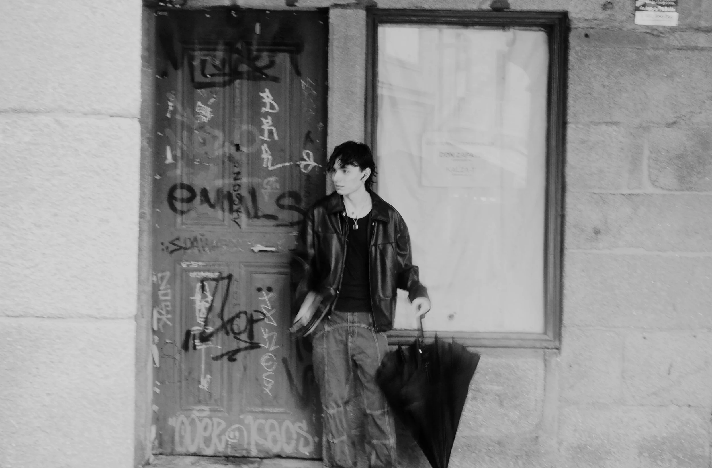 A young person with short dark hair wearing a black leather jacket, black shirt, and loose jeans stands against a graffiti-covered wooden door holding a black umbrella.
