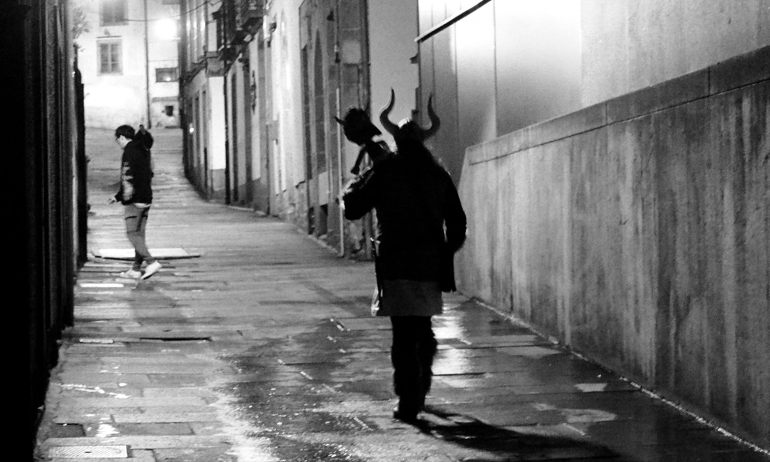 A person dressed in a costume with horns and a dark outfit walking on a city sidewalk at night, with another person looking at their phone in the background.