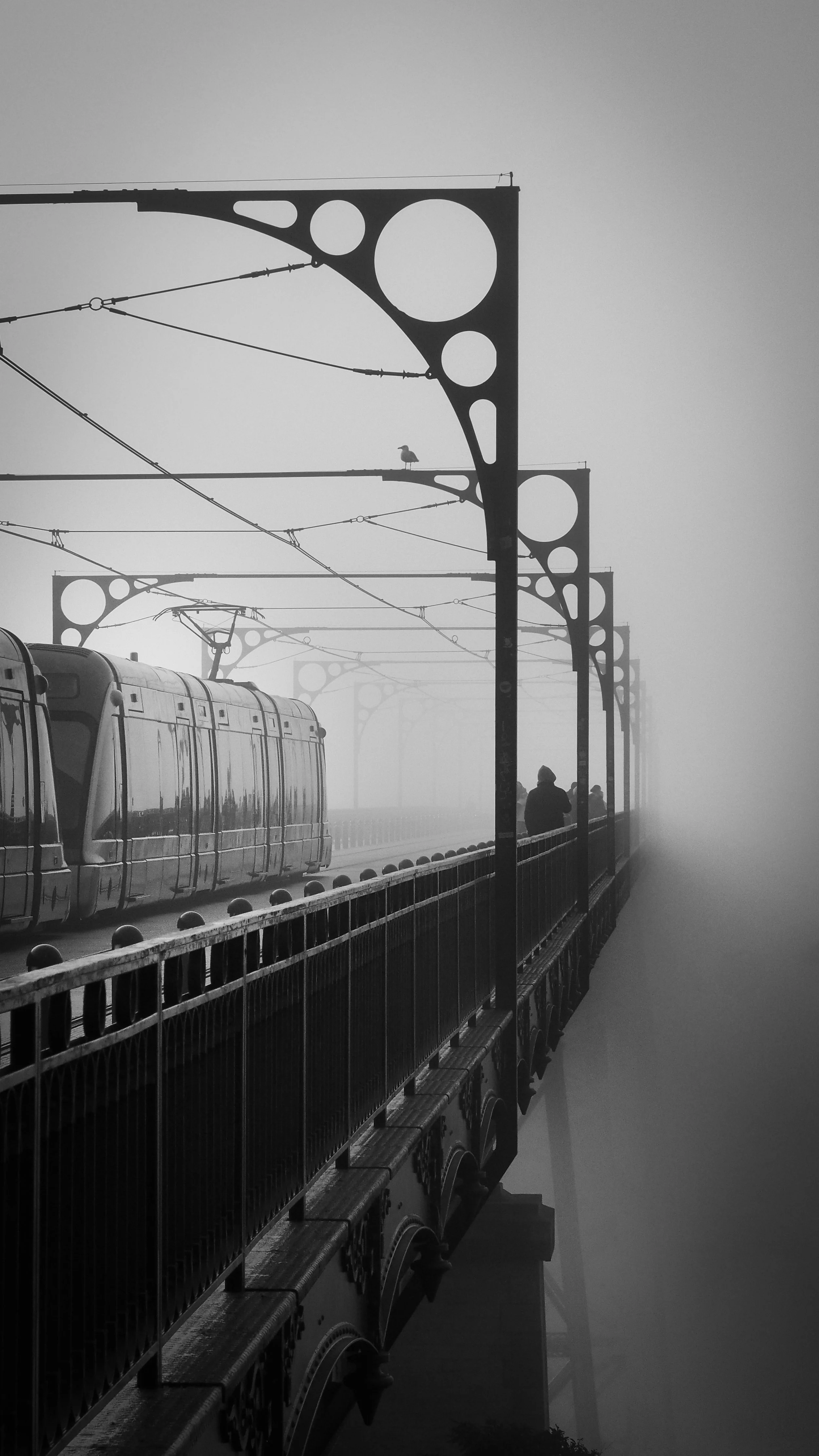 Black and white photo of a foggy bridge with a train passing and a person walking on the side, with decorative metal arch supports.