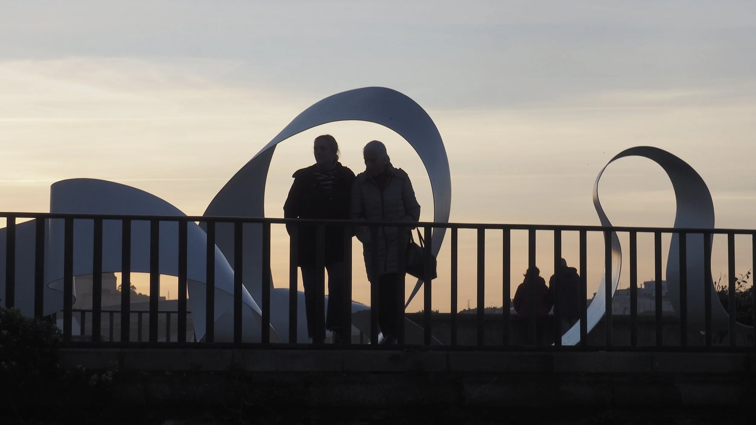 Silhouettes of two people walking on a bridge with modern, abstract sculptures behind them, at sunset.