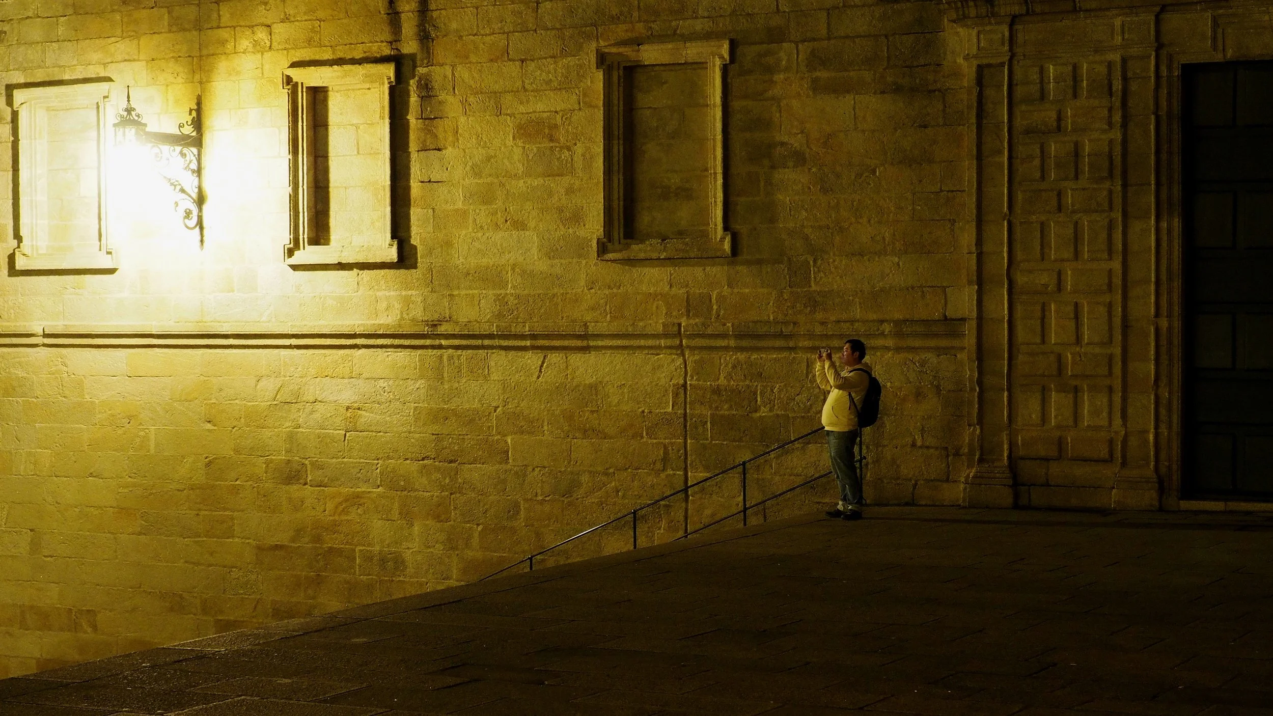 A person standing on a slightly elevated surface, taking a photograph or video with a small device, next to a large stone wall illuminated by a yellowish light at night.