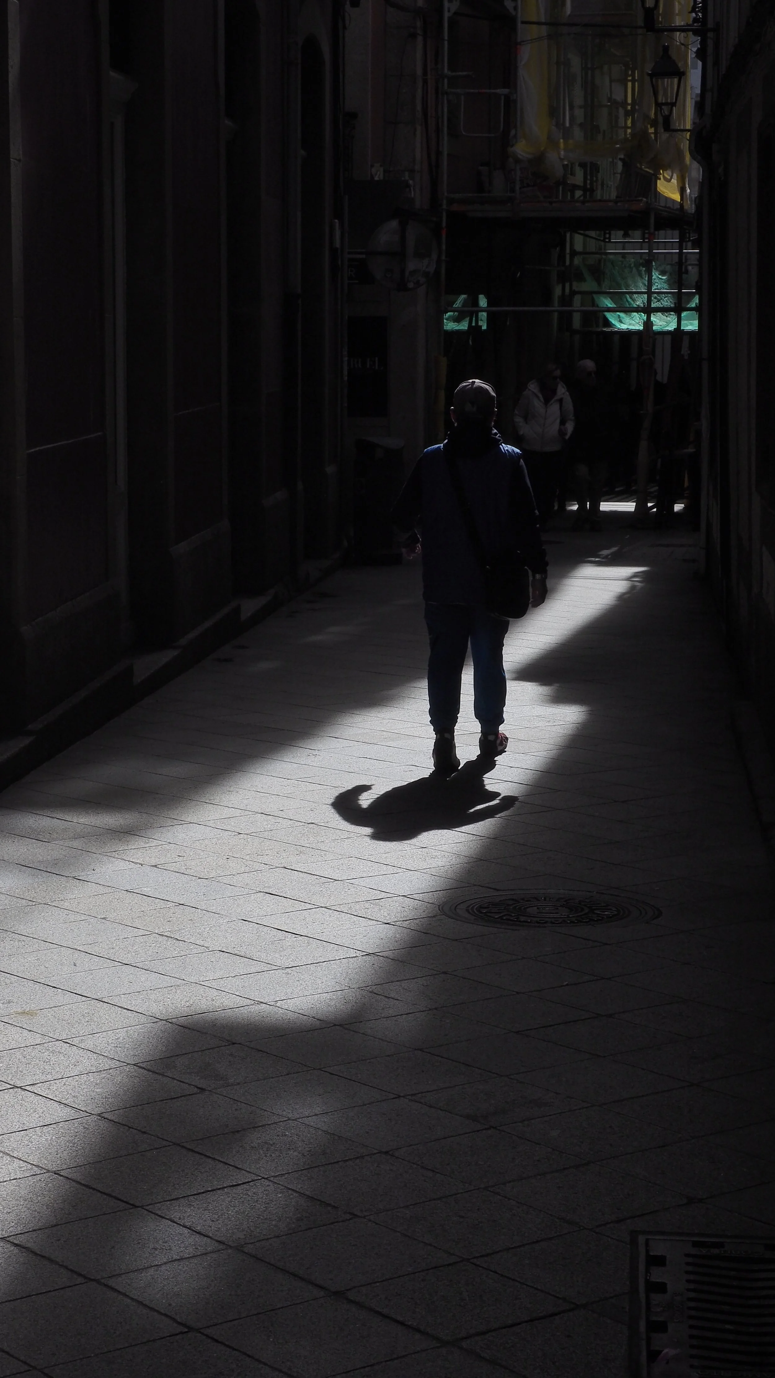 A person walking in a dark alleyway with bright sunlight creating a shadow on the ground, and a few people in the background.