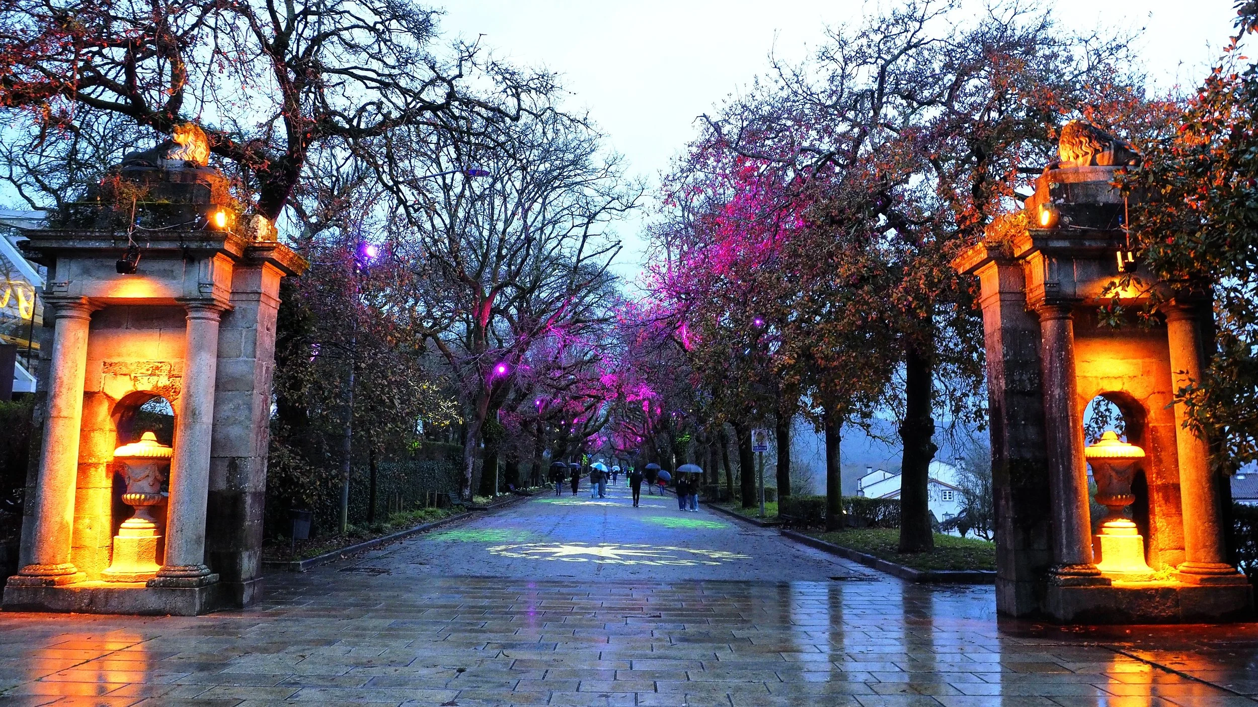 A wet, stone-paved pathway lined with trees at dusk, with pink and purple blossoms and decorative lighting. Stone columns with urns and fires flank the entrance.