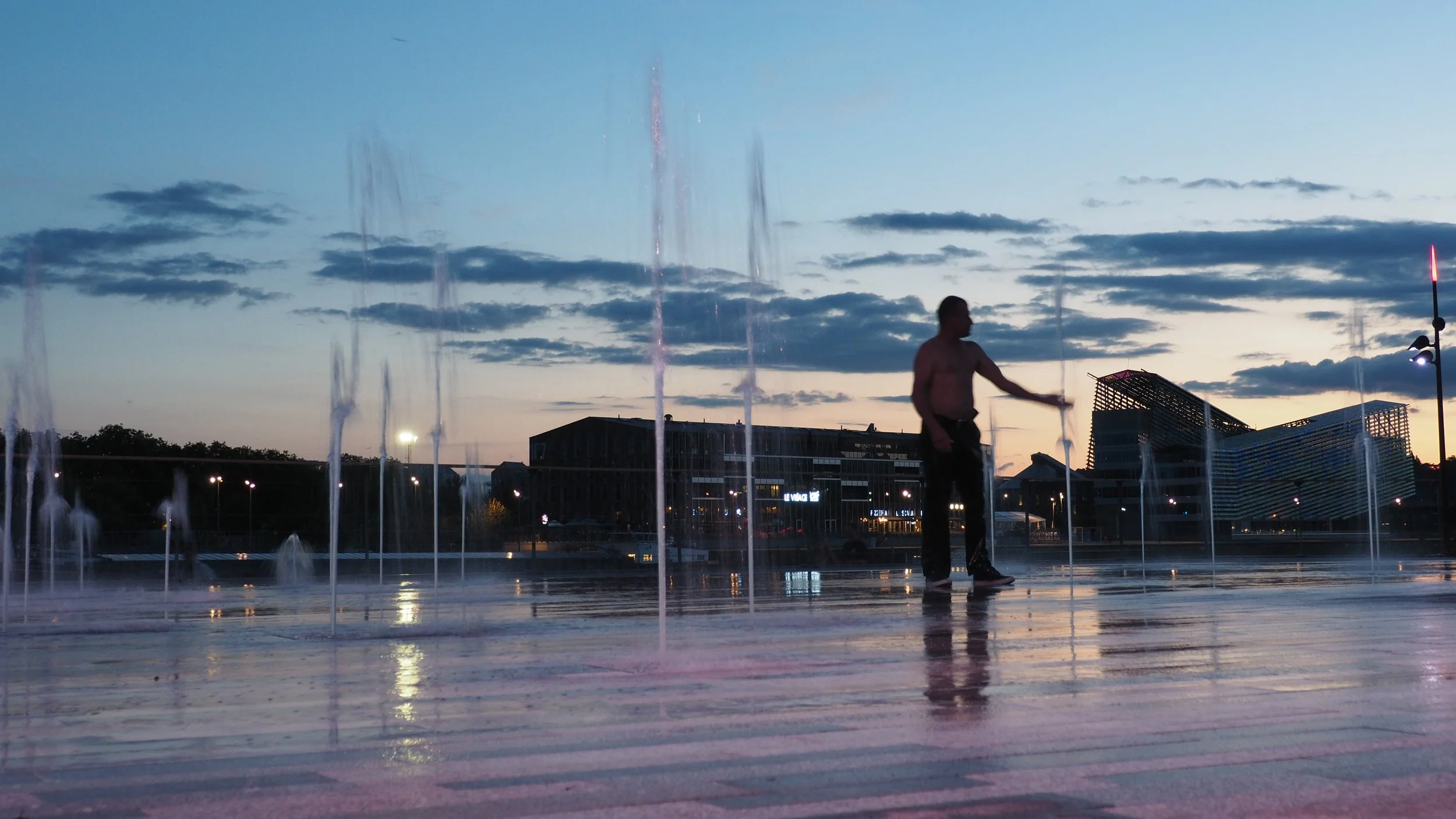 A man in dark pants is playing with water jets on a public fountain during dusk, with modern buildings and a partly cloudy sky in the background.