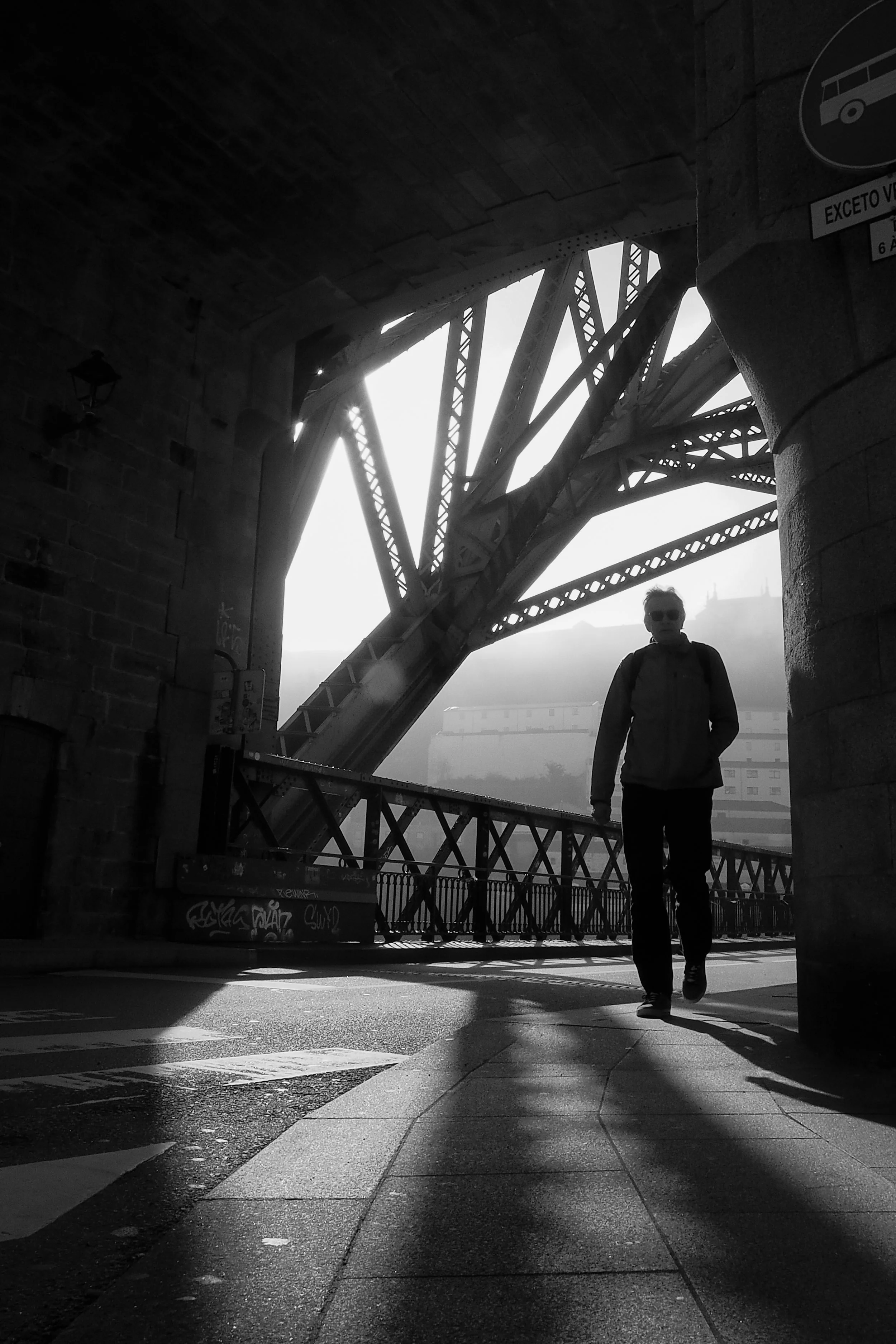 A man walking under a bridge with steel beams, casting a shadow on the pavement, in black and white.