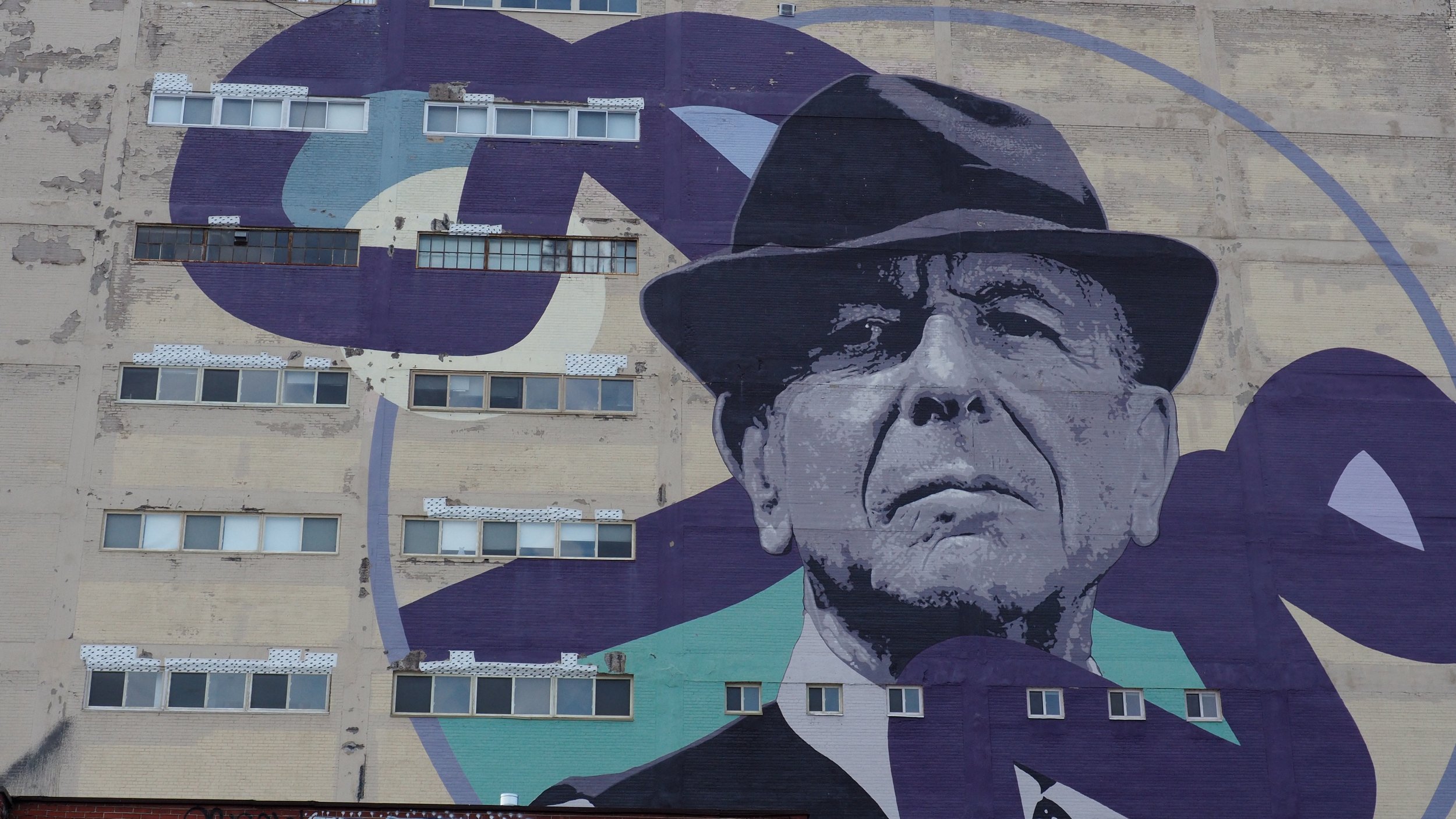 A mural of an elderly man wearing a hat painted on the side of a building with multiple windows, using shades of purple, black, and white.