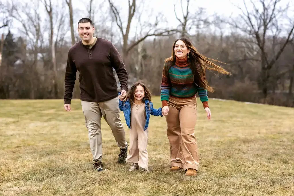 A family of three, a man, a woman, and a young girl, holding hands and walking across a grassy field outdoors on a cloudy day, smiling and enjoying each other's company.