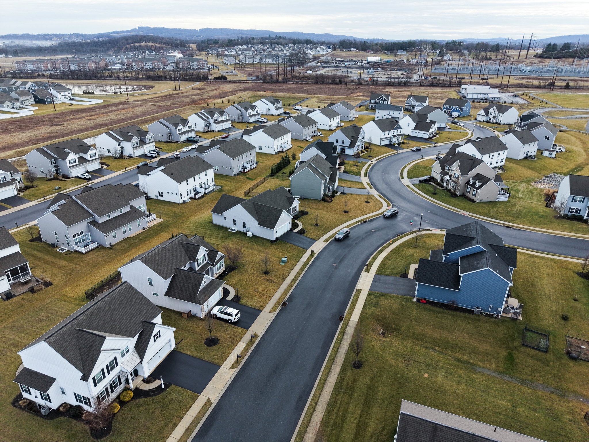 A neighborhood with multiple single-family houses with yards and driveways, curving paved roads, and a background of open fields, power lines, and a cloudy sky.
