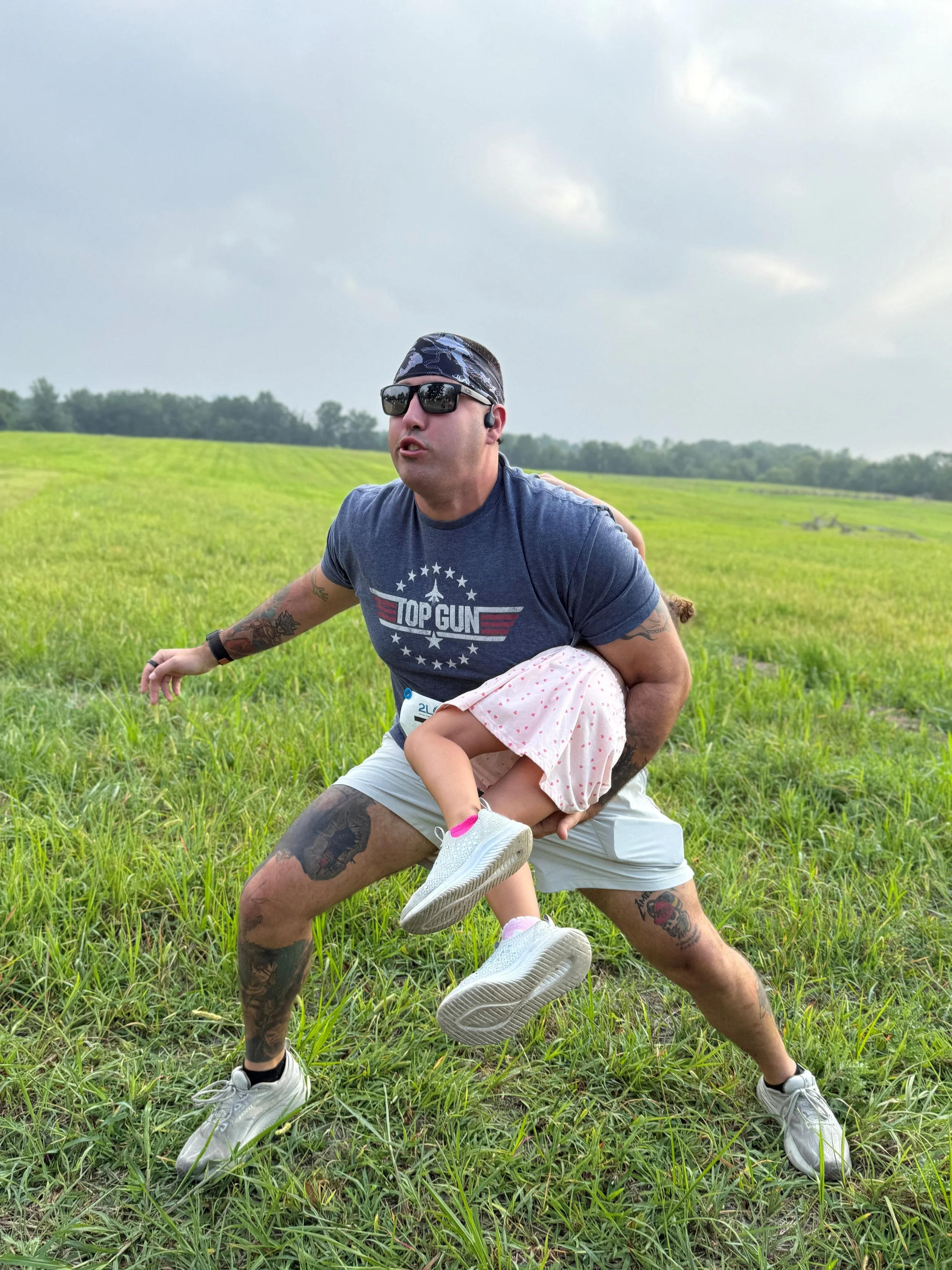A man wearing sunglasses, a bandana, and a blue T-shirt with the text "Top Gun" is holding a young girl in a pink polka dot dress with white sneakers, in a grassy outdoor field under cloudy skies.