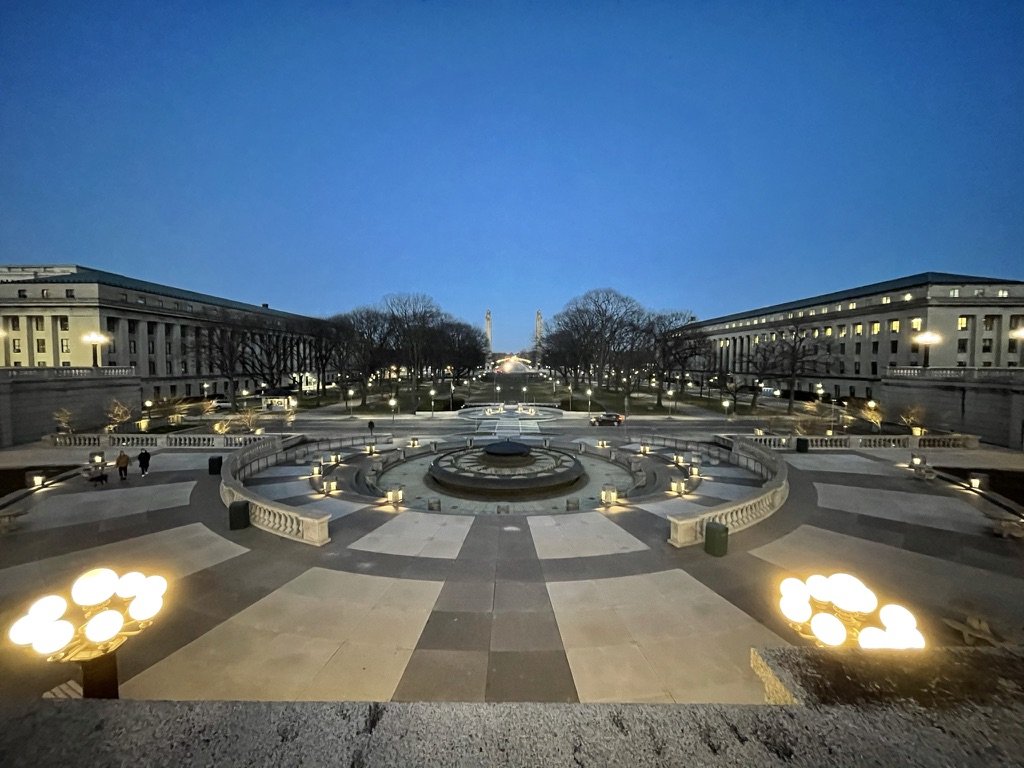 A city square at dusk with large buildings, trees, a monument, and a wide open courtyard, illuminated by streetlights and decorative lamps.