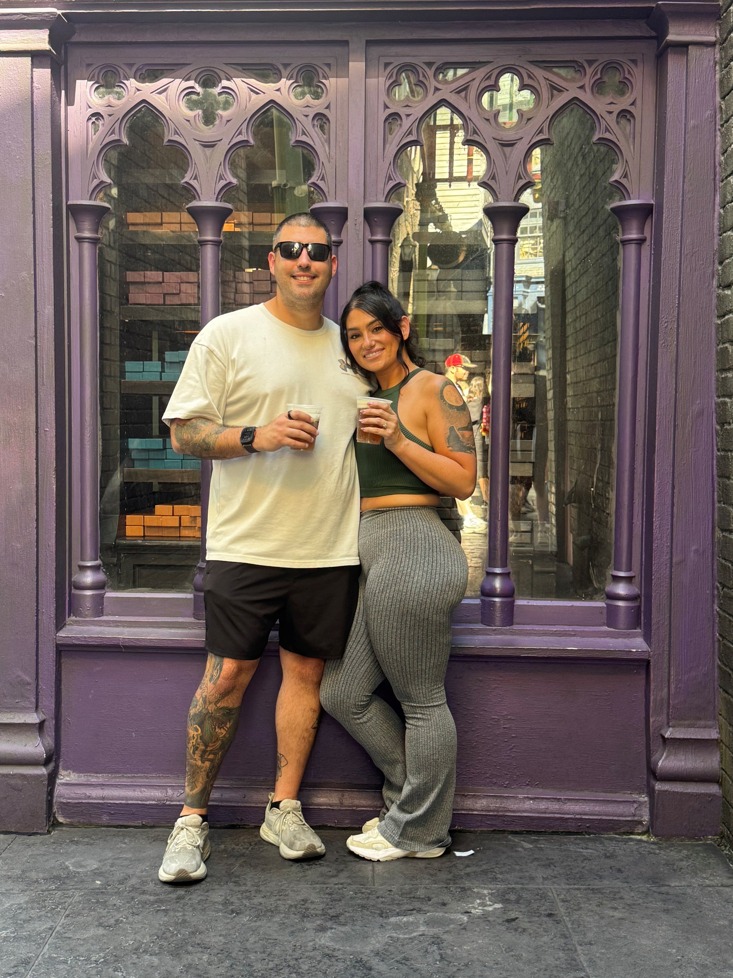 A man and woman stand together outside a decorative purple window, each holding a drink and smiling at the camera.