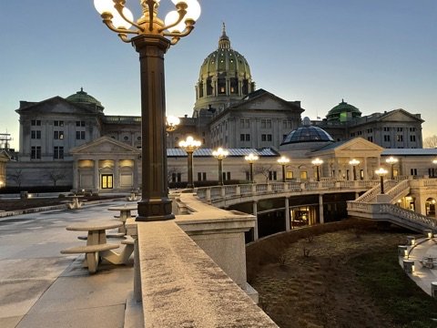 View of a historic government building with a green domed roof, seen from a terrace with decorative street lamps and picnic tables during dusk.