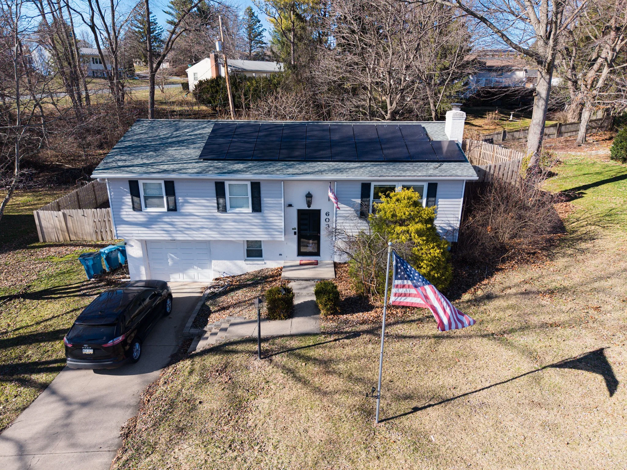 Aerial view of a white suburban house with black shutters, a driveway with a parked black car, and solar panels on the roof. An American flag is in front of the house, with a small front yard and a large yard with trees and a wooden fence in the background.