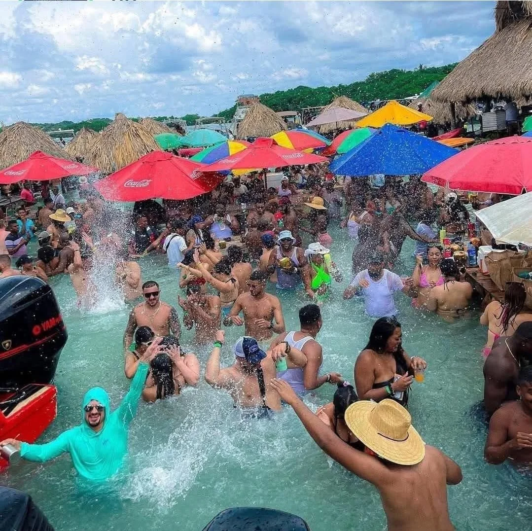 Muchas personas disfrutando en una piscina con sombrillas y toldos en un ambiente tropical y festivo.