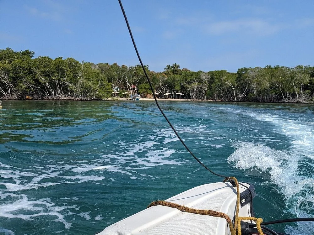 Imagen de un bote navegando por un río rodeado de árboles verdes en una zona tropical, con una cuerda atada en la parte delantera del bote y la moto de agua dejando estelas en el agua.