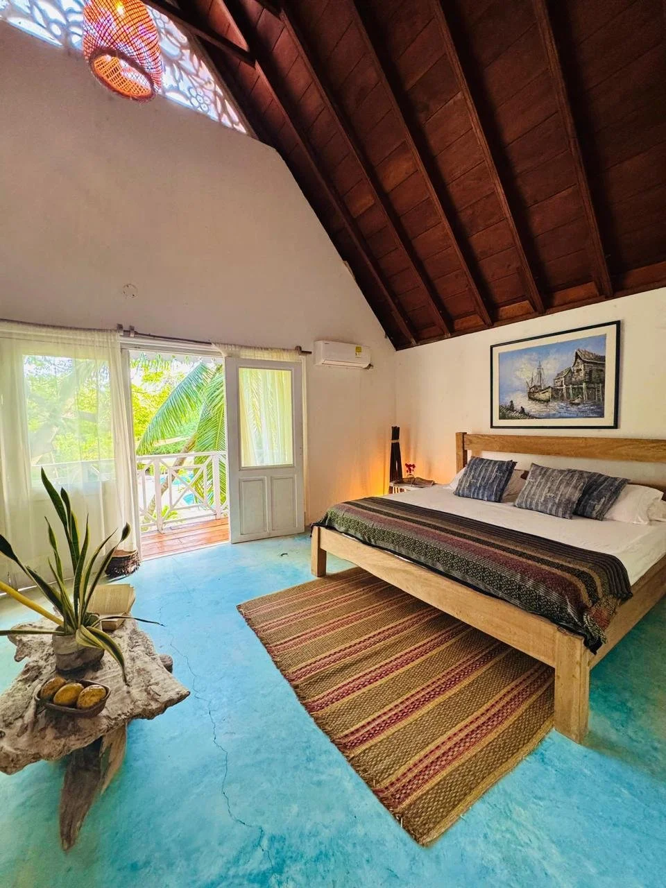 Habitación con un cómodo cama de madera, alfombra tejida, ventana con vista a vegetación tropical, y techo de madera en forma de catedral.