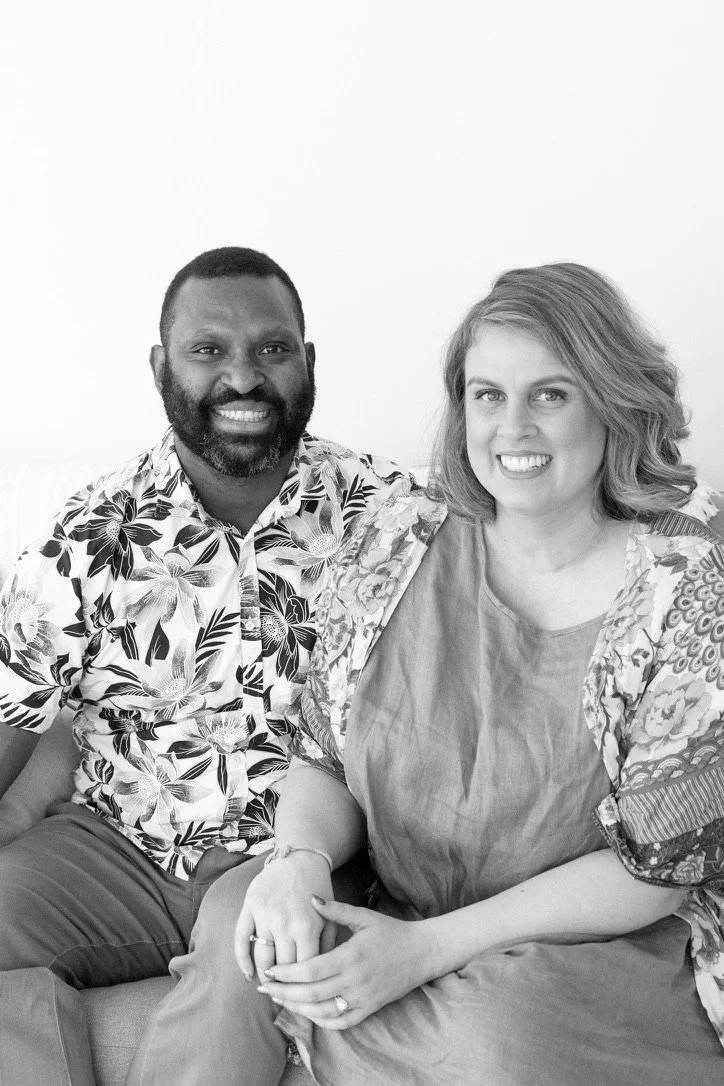 A smiling black man with a beard and a woman with shoulder-length hair sitting close together on a couch, holding hands, in a well-lit indoor setting.