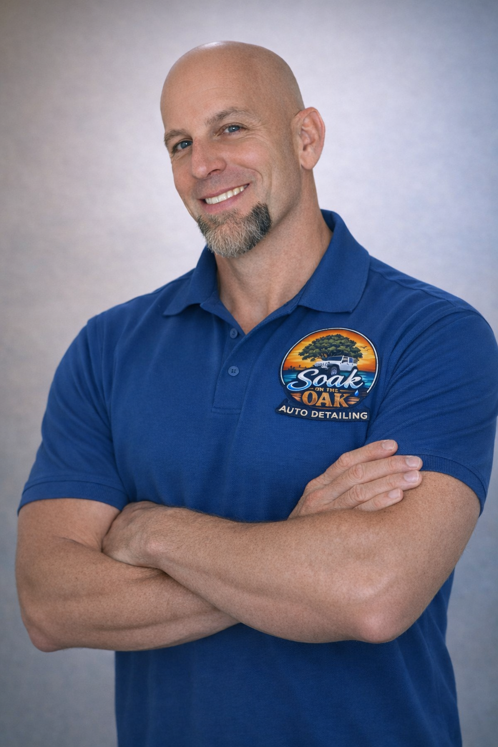Owner wearing a blue polo shirt with an "Soak on the Oak Auto Detailing" logo, standing against a neutral background with arms crossed and smiling.