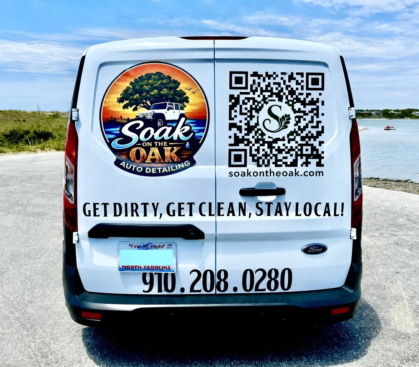 Mobile detailing white van with a logo of a tree, car, sunset, and water reading 'Soak on the Oak Auto Detailing' parked near water and grassy area, with a QR code and the tagline 'Get dirty, get clean, stay local!'. 