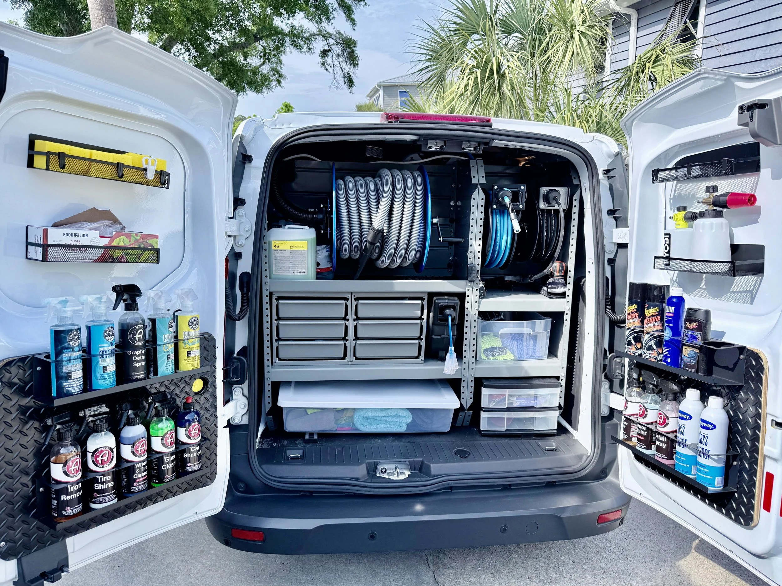 Open back of a white mobile detailing van filled with cleaning supplies, including spray bottles, containers, and organizing bins.