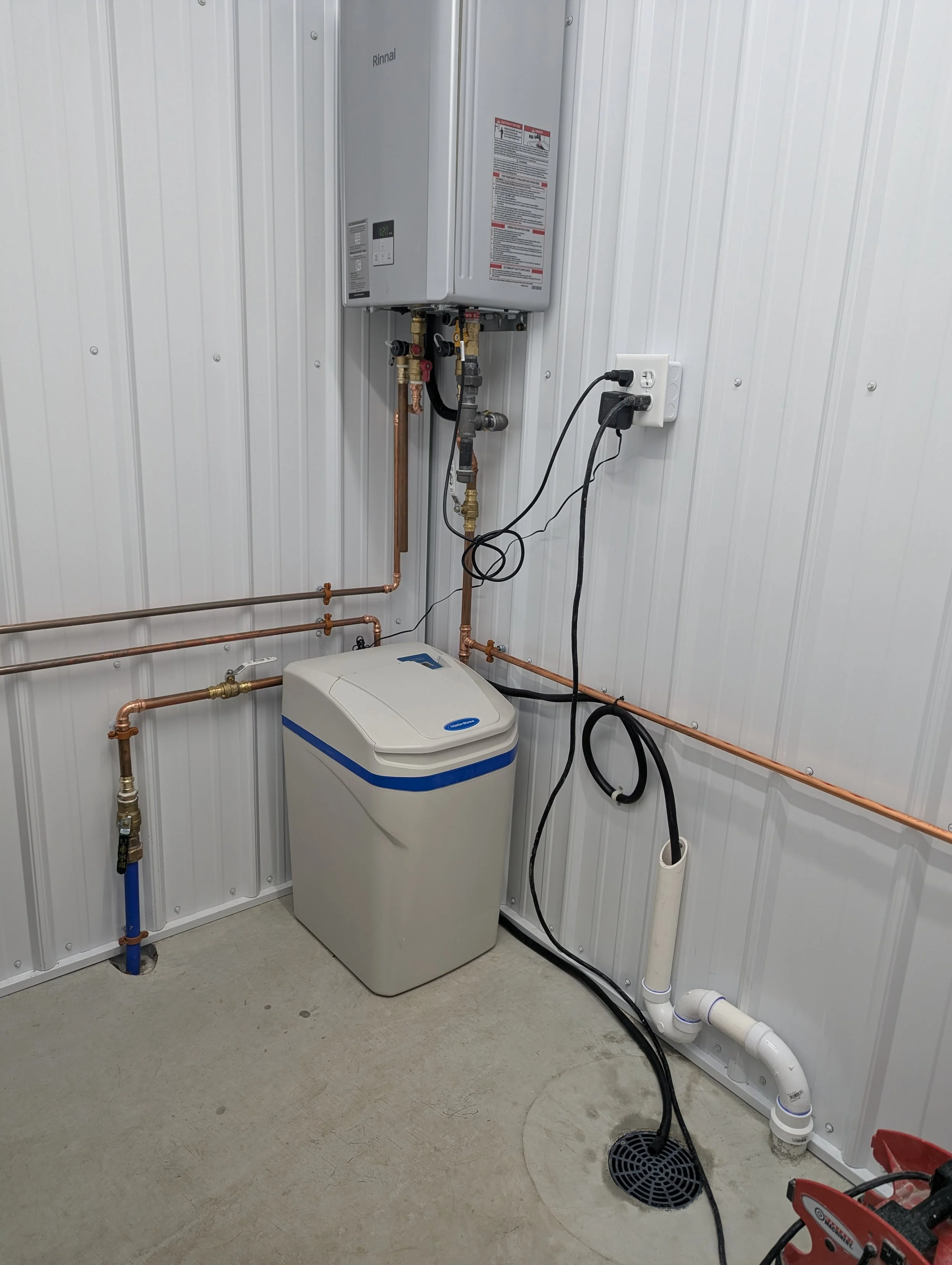 A utility room with a Rinnai tankless water heater mounted on the wall, copper pipes, a white water softener, electrical cords, and a floor drain.