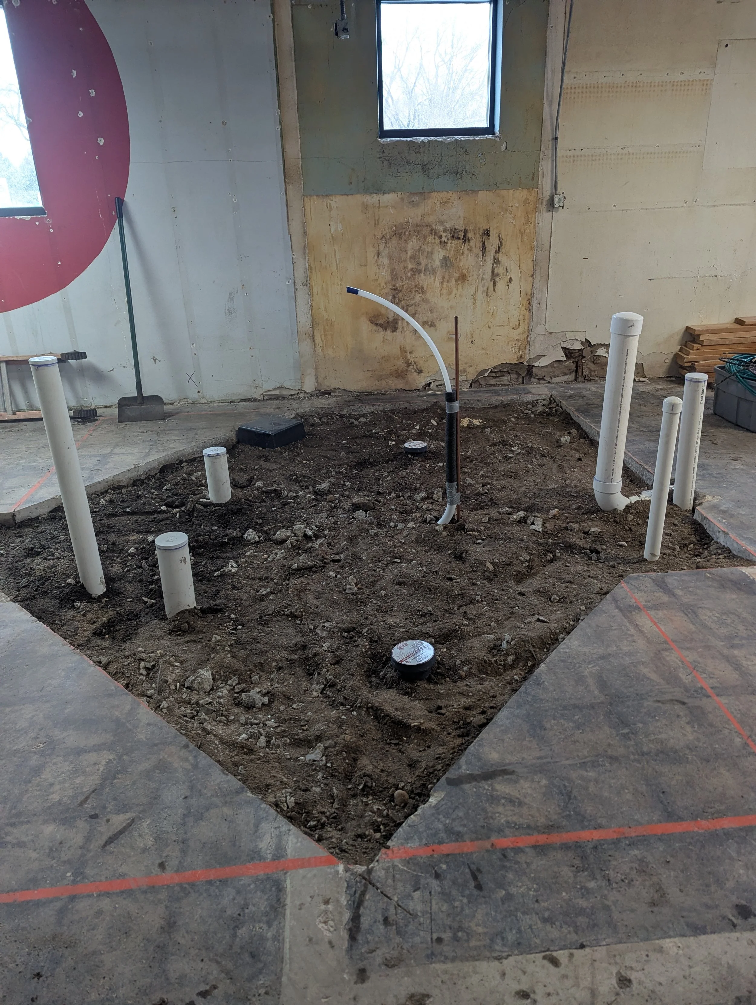 Interior of a building under construction with exposed soil, several white plumbing pipes, and a curved pipe with a flexible hose, surrounded by unfinished walls and construction tools.