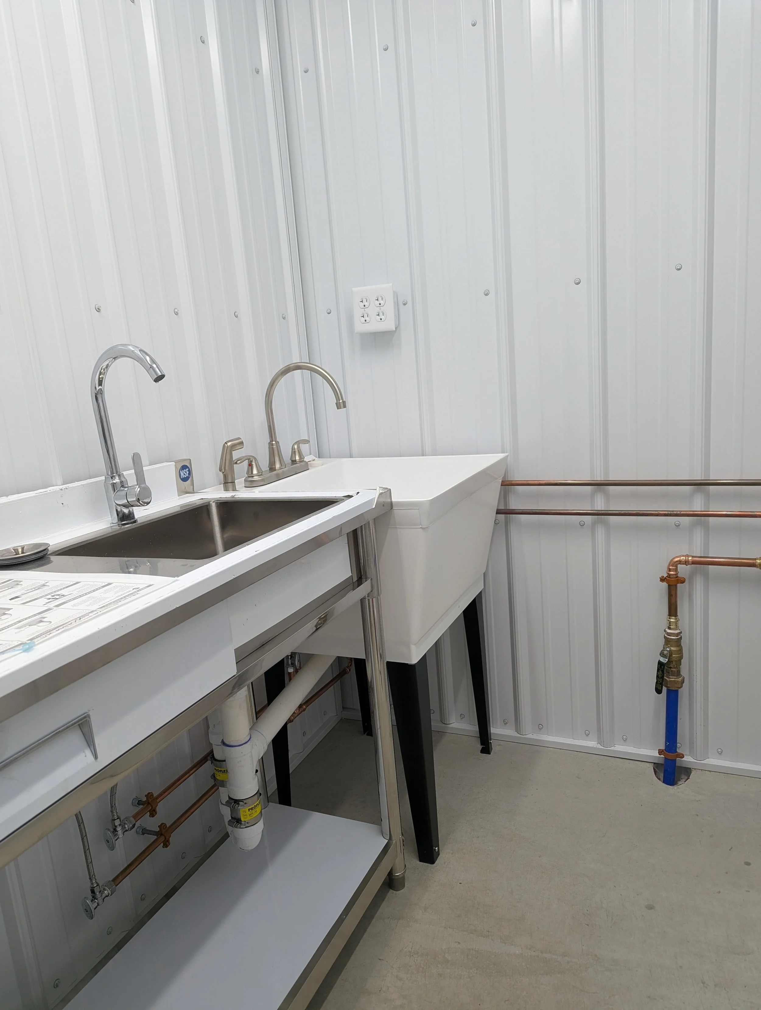 A utility room with white panel walls, a stainless steel sink with two faucets, a white hand washing sink, a wall outlet, and exposed copper piping.