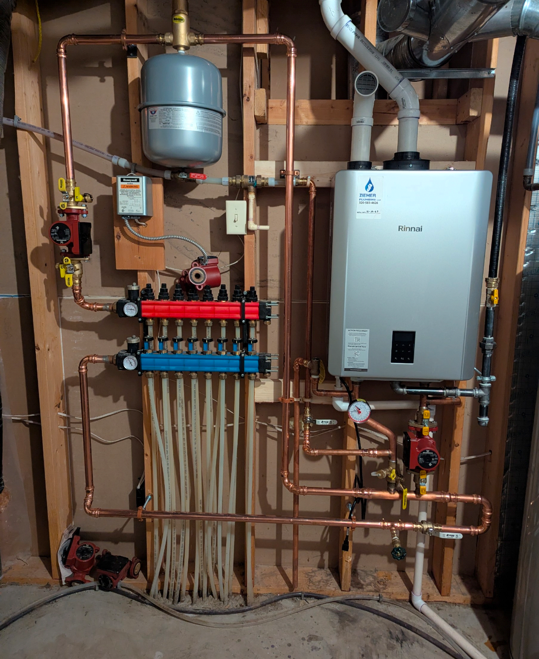 A plumbing and heating system with copper pipes, a water heater, a manifold with red and blue valves, and various gauges and control components installed on a wooden wall framing.