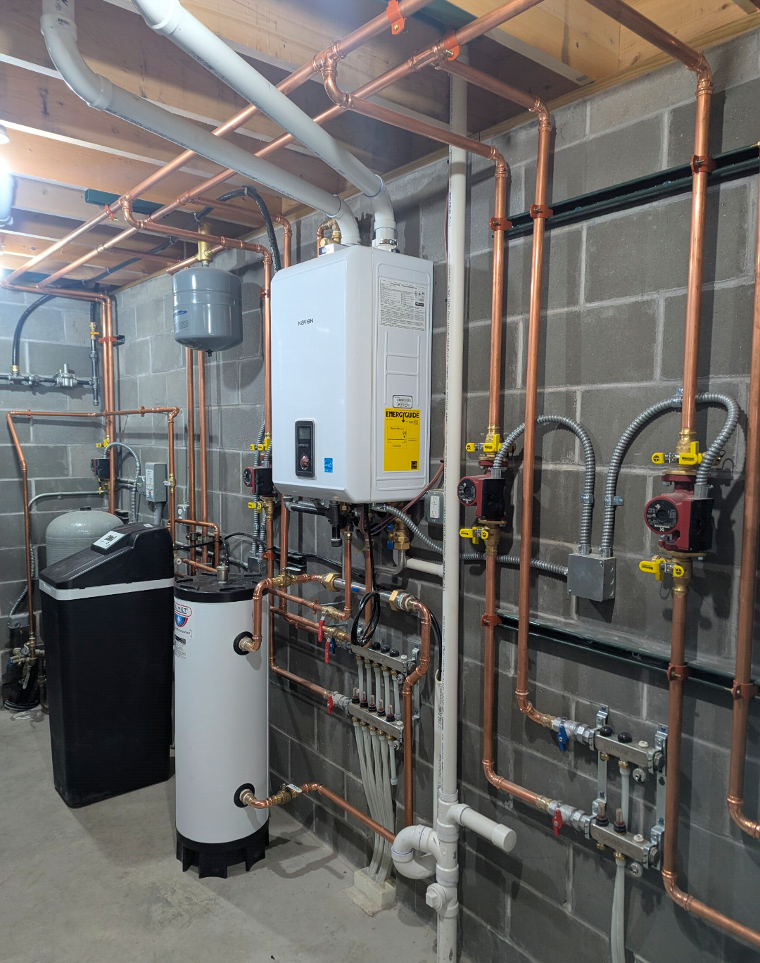 A utility room with copper and PVC pipes, a white Navien tankless water heater, a water softener, and a gray expansion tank on a concrete floor with unfinished cinder block walls.