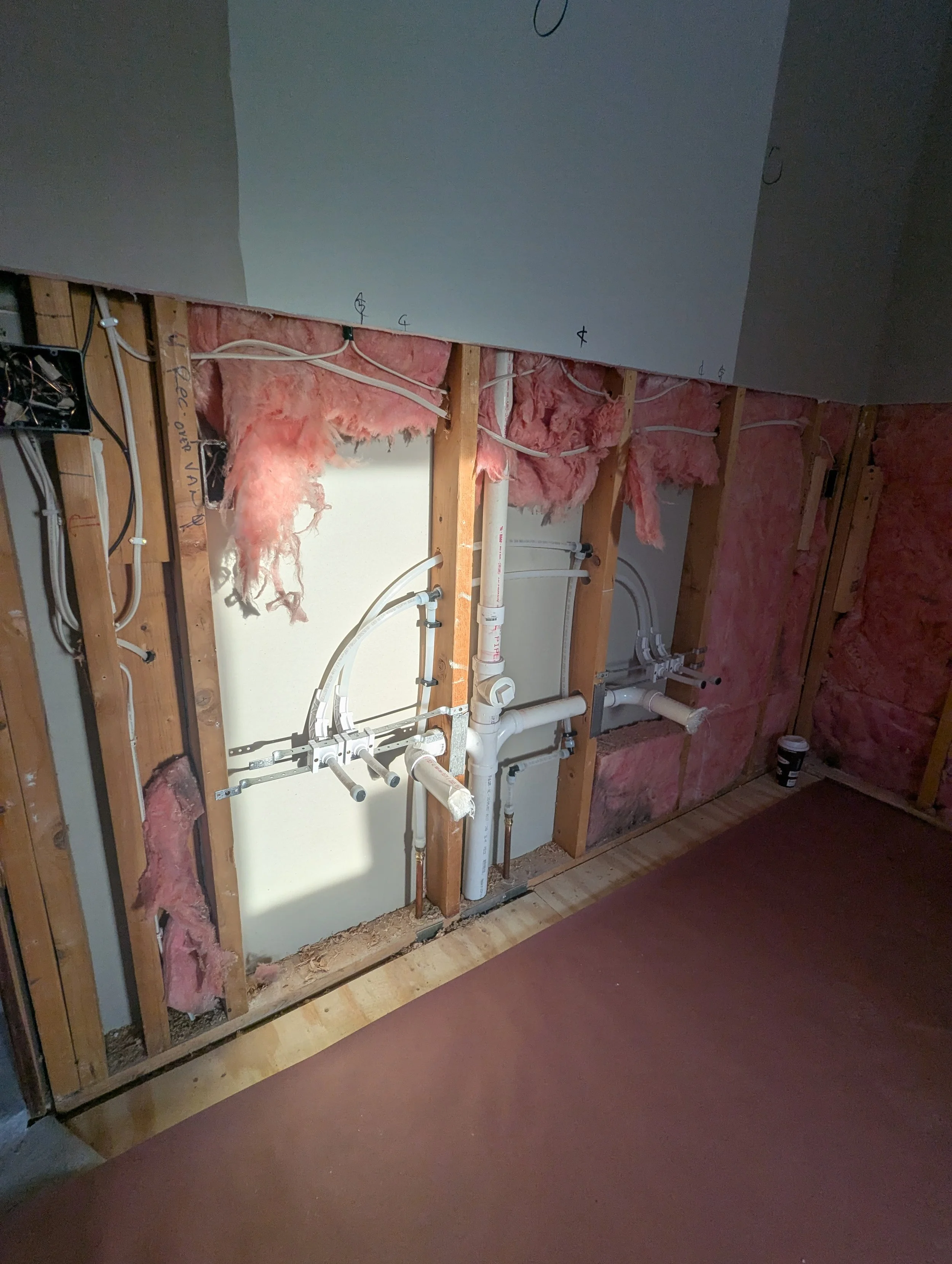 Partially finished wall in construction with exposed pink insulation, plumbing pipes, and electrical wiring.