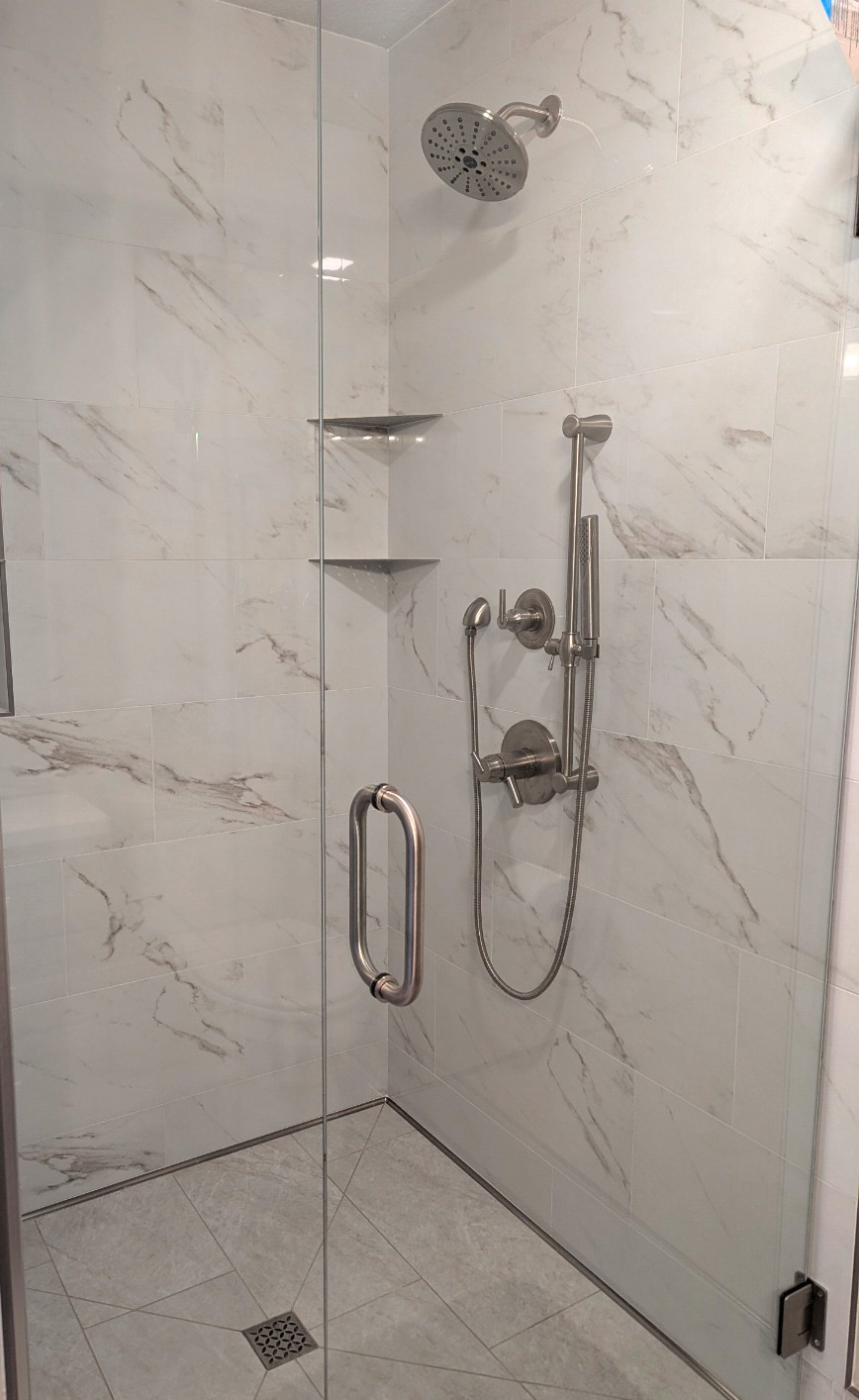A modern walk-in shower with marble tile walls, a rainfall showerhead, a handheld shower, and built-in corner shelves, enclosed by a glass door.